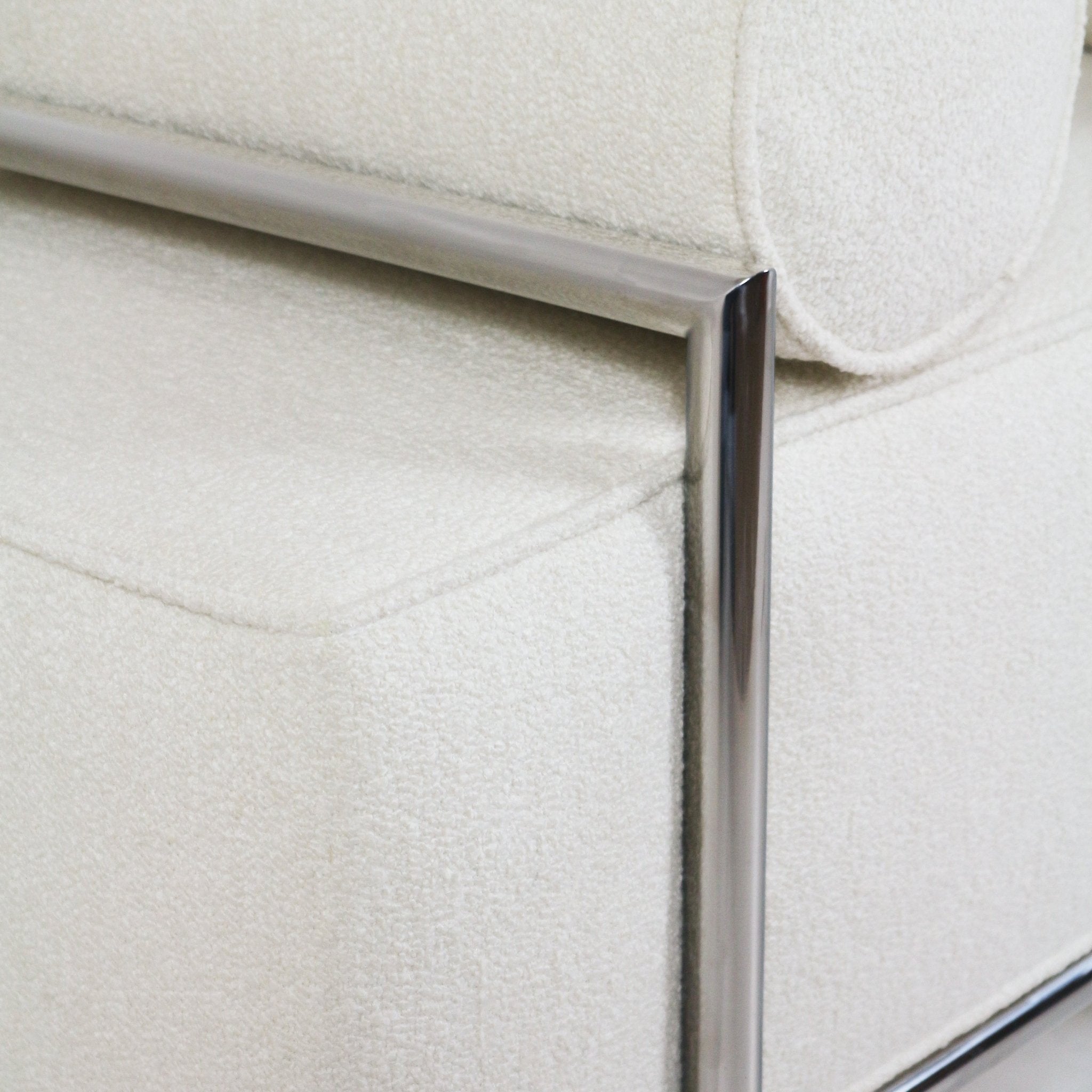 Close-up of a white boucle-upholstered end-of-bed bench showing textured fabric, stitched seat seam, round bolster cushion, and polished stainless steel frame.