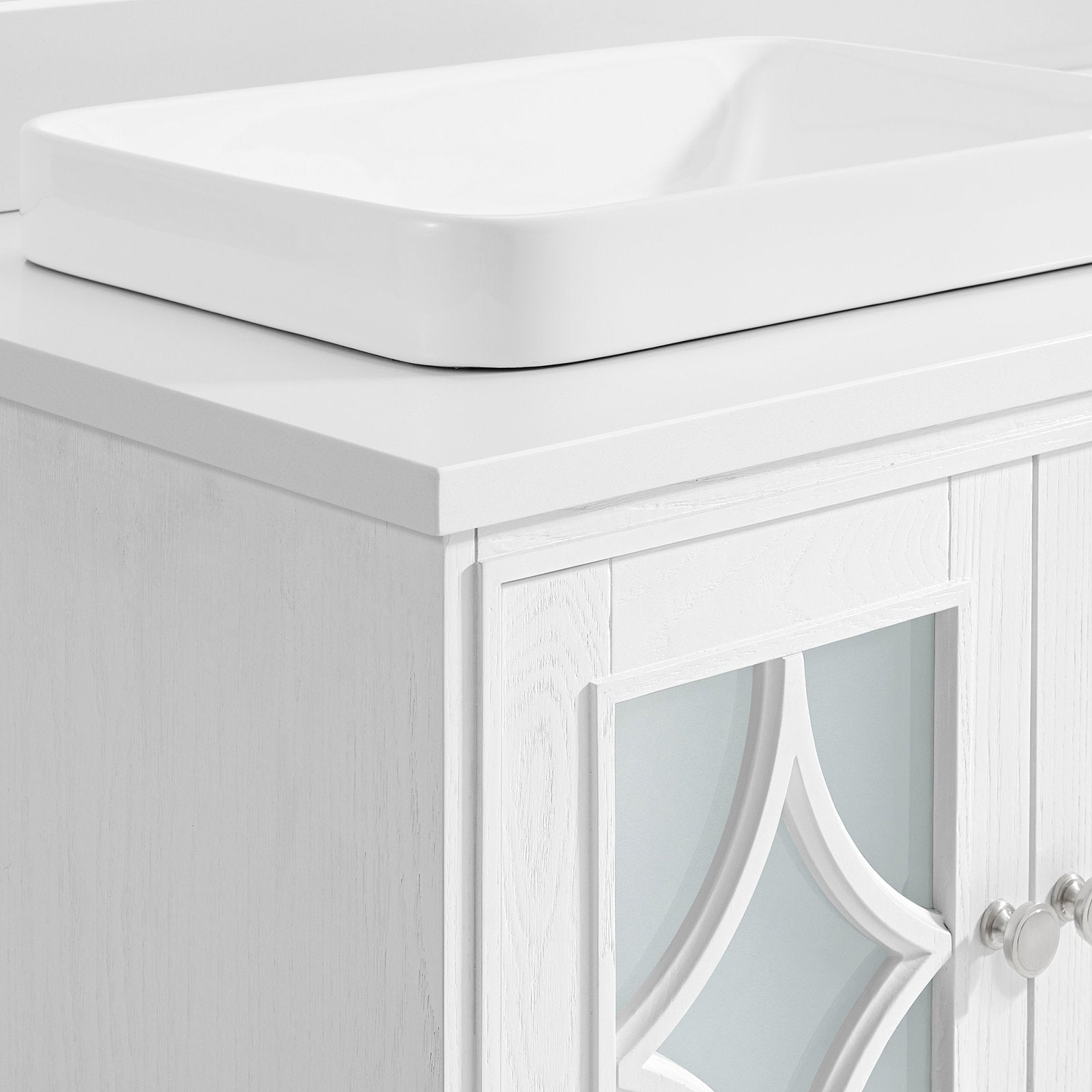 Close-up of a white ceramic rectangular vessel sink on a stone top above a painted solid wood vanity with a decorative frosted-glass door panel and round metal knobs.