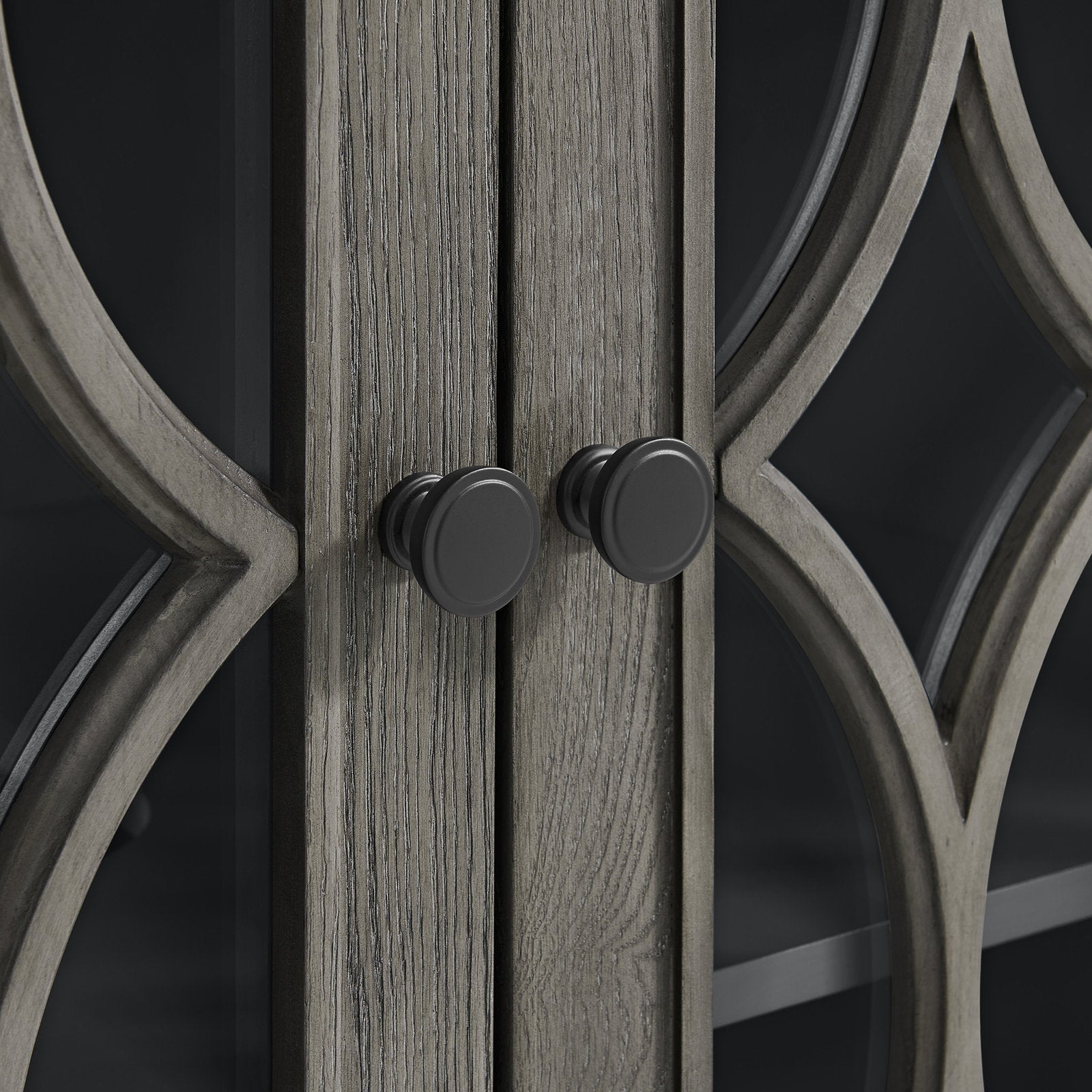 Close-up of stone-colored solid wood vanity doors with visible wood grain, decorative curved wood mullions over glass panels, and two matte black round cabinet knobs.