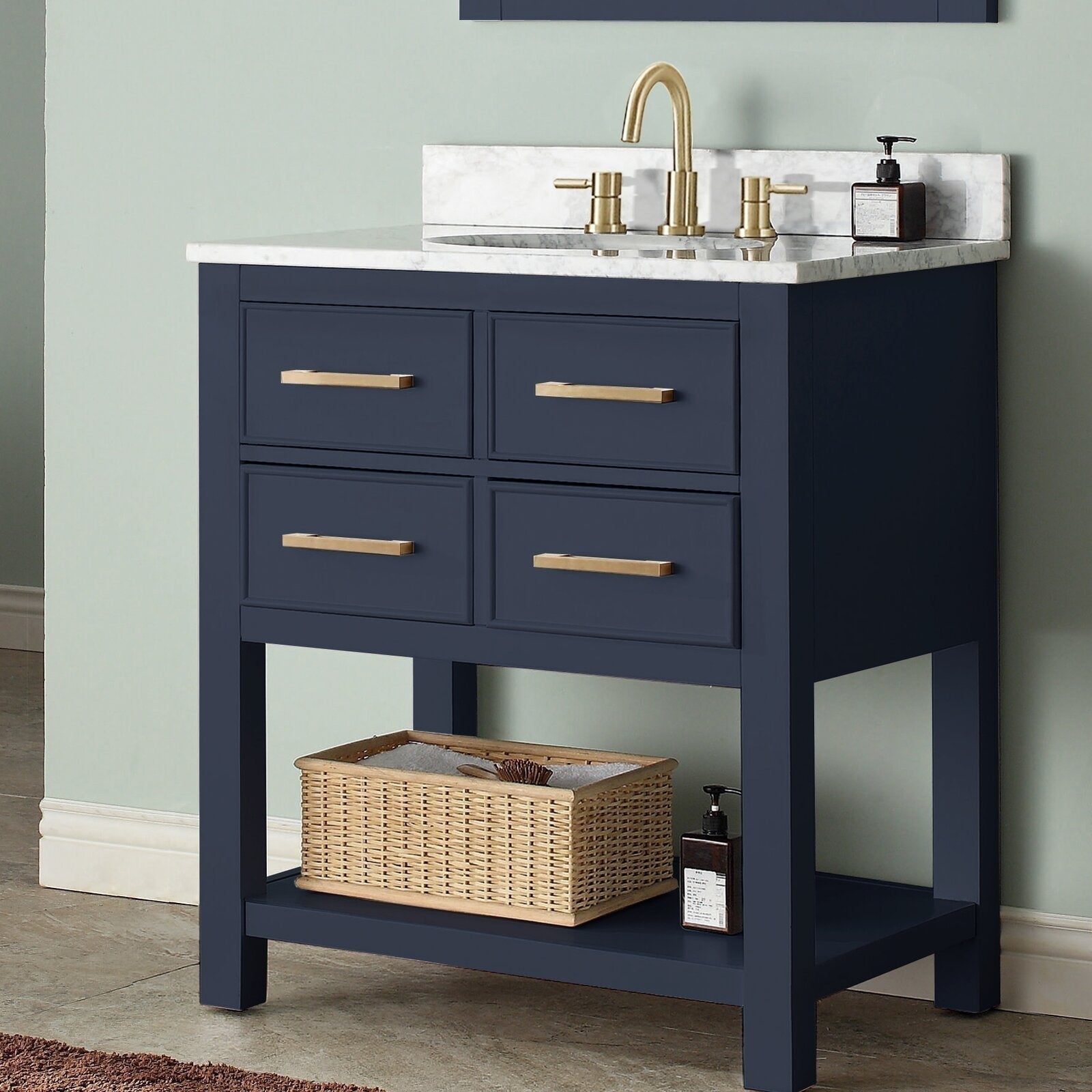 Freestanding modern solid-wood bathroom vanity with a stone countertop and ceramic undermount sink, navy painted wooden base with four drawers, brass bar pulls and an open lower shelf holding a wicker basket and soap dispenser.