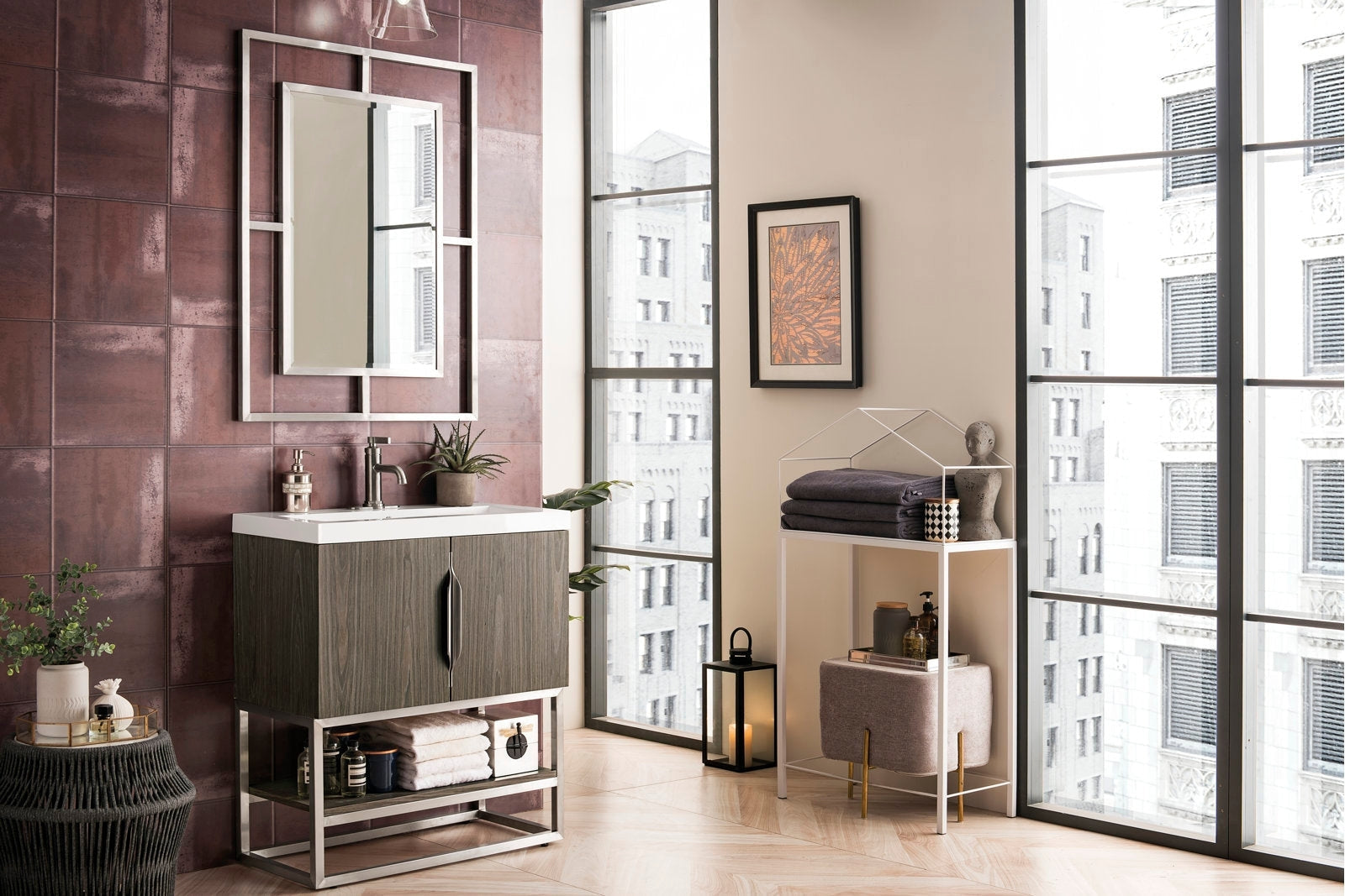 Freestanding modern single-sink bathroom vanity with a white stone top, solid wood stone-finish cabinet on a chrome metal base, framed mirror, burgundy tile wall and styled towels and plants.