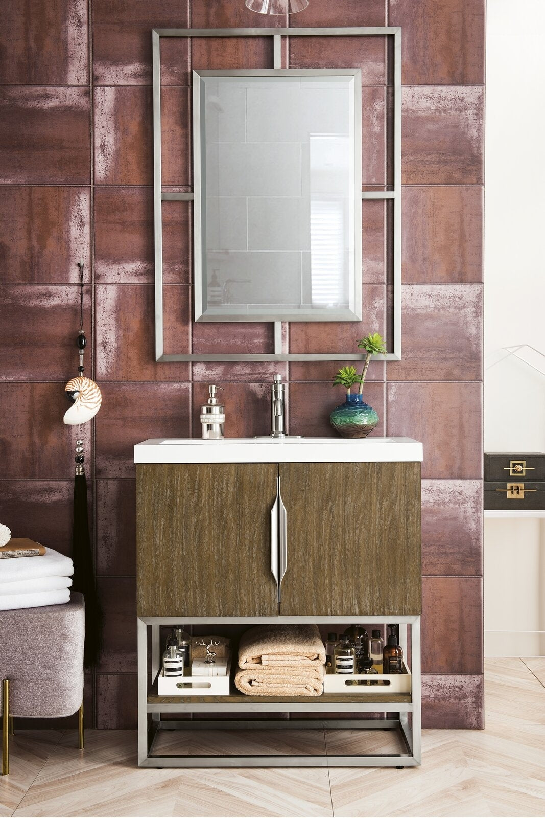 Modern solid wood bathroom vanity with a white stone top, chrome faucet and integrated sink, vertical metal handles, open lower shelf holding folded towels and bottles, framed rectangular mirror and rust-red tiled wall backdrop.