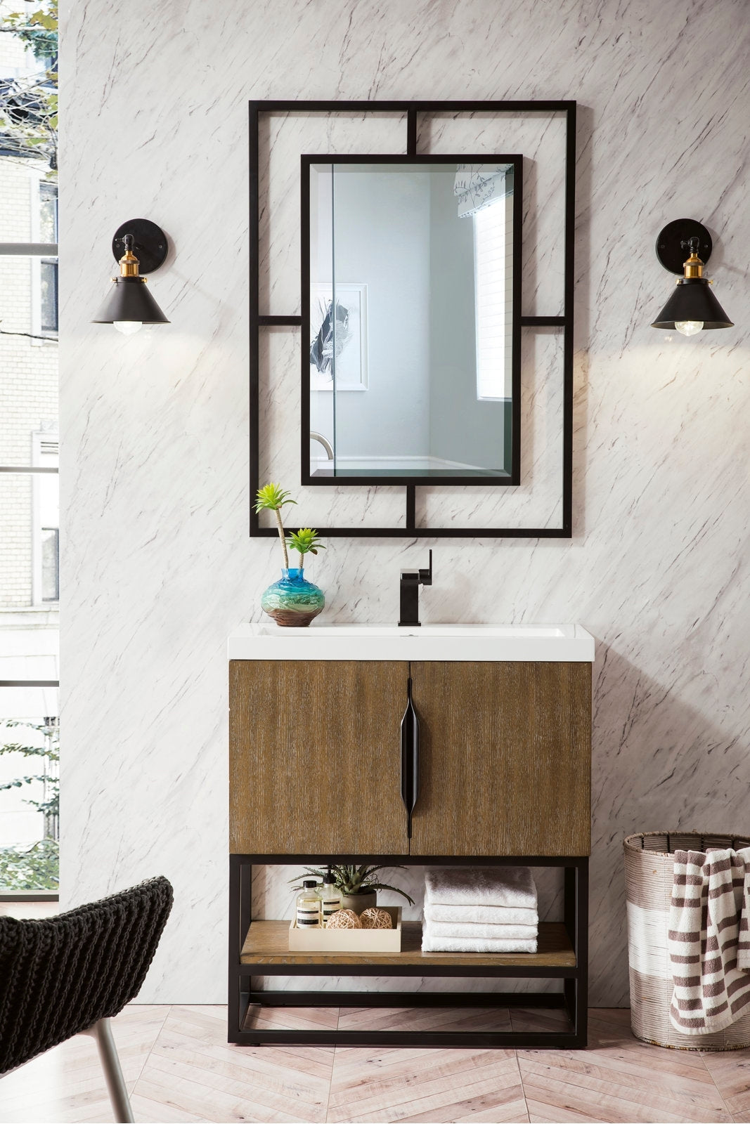 Freestanding modern single-sink bathroom vanity with a white stone top and stone-colored solid wood cabinet, black metal base and geometric framed mirror, black faucet, twin wall sconces and an open lower shelf with towels.