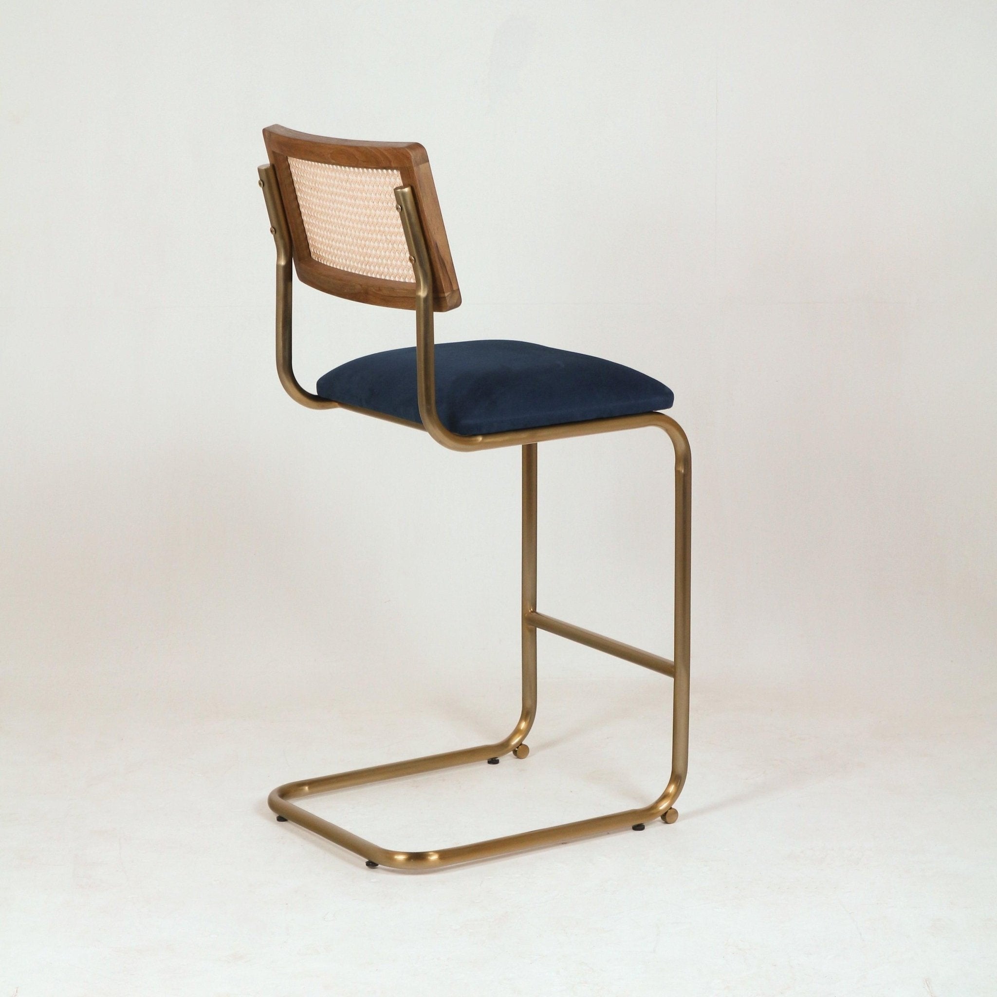Cantilever counter-height bar stool with a navy velvet upholstered seat, curved brass-finish metal frame and a wood back with woven cane panel, shown on a plain white studio background.