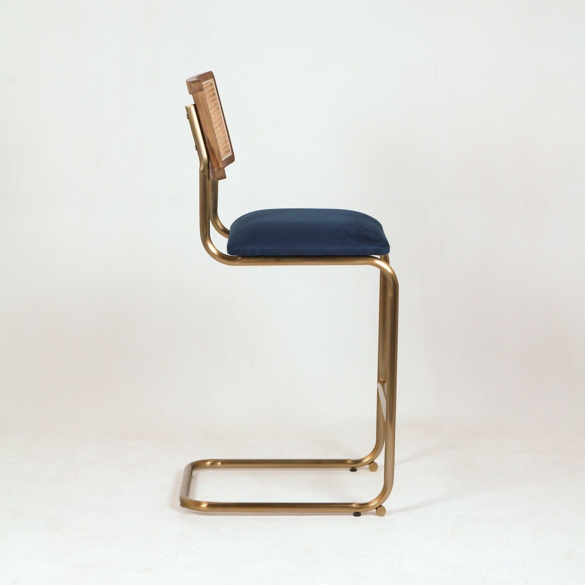 Side view of a modern cantilever bar stool with a brass metal frame, navy velvet seat cushion and wood-framed woven cane back against a white background.