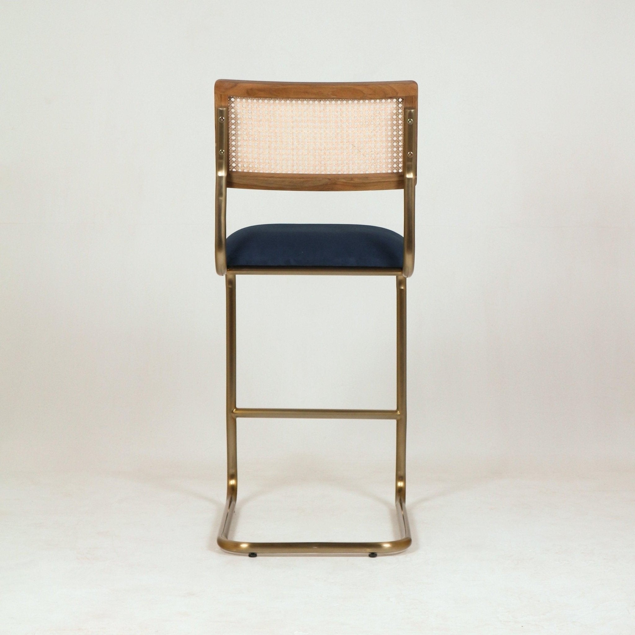 Rear view of a modern cantilever bar stool with a brass metal frame, navy blue upholstered fabric seat, natural cane back panel and walnut wood top rail against a neutral studio background.