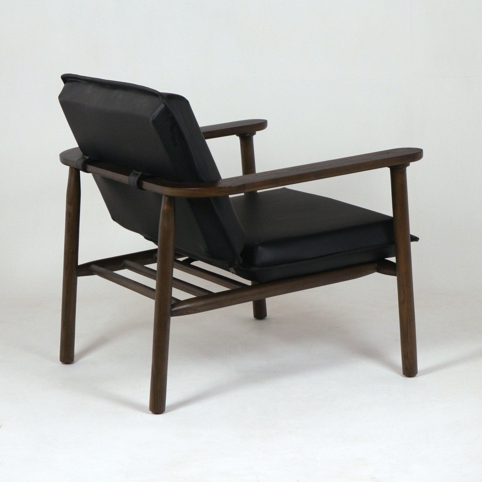 Rear three-quarter view of a midcentury lounge chair featuring a tapered solid wood frame and arms, black faux leather seat and back cushions, and a slatted wooden support shelf beneath the seat.