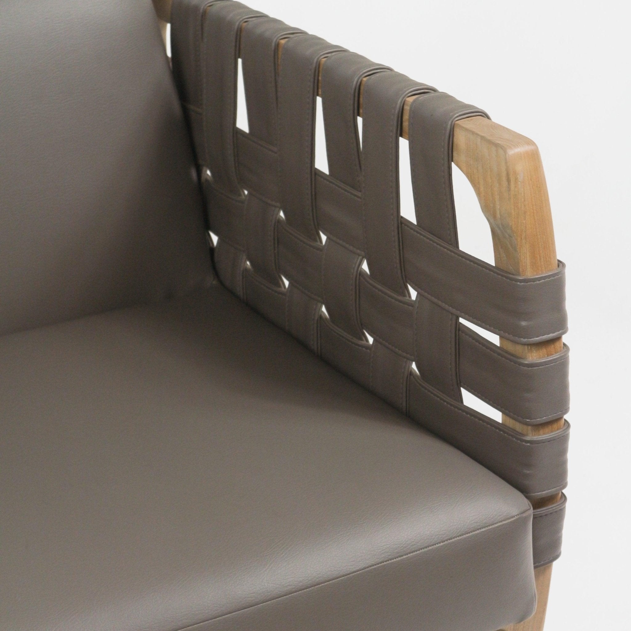 Close-up of a modern lounge chair showing a brown faux-leather seat cushion and woven webbed armrest of wide leather straps wrapped around a natural solid wood frame.