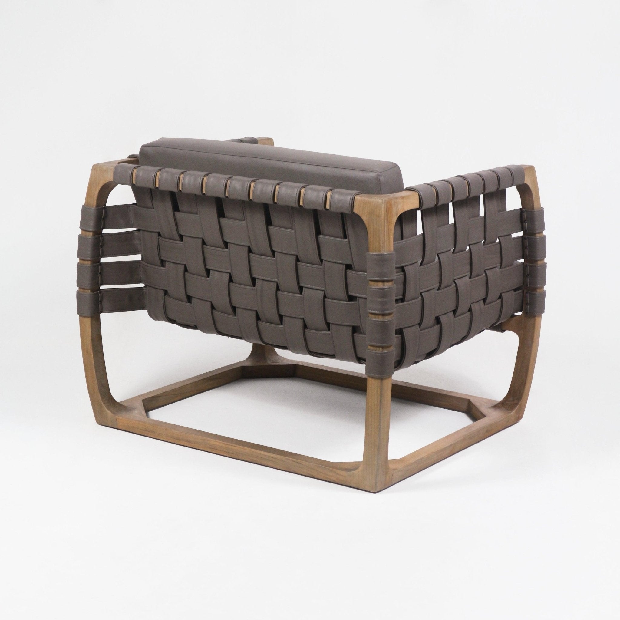 Rear three-quarter view of a modern low-profile lounge chair with a solid natural wood frame and wide dark brown faux-leather woven straps forming the seat and back, topped by a matching cushioned low back pillow.
