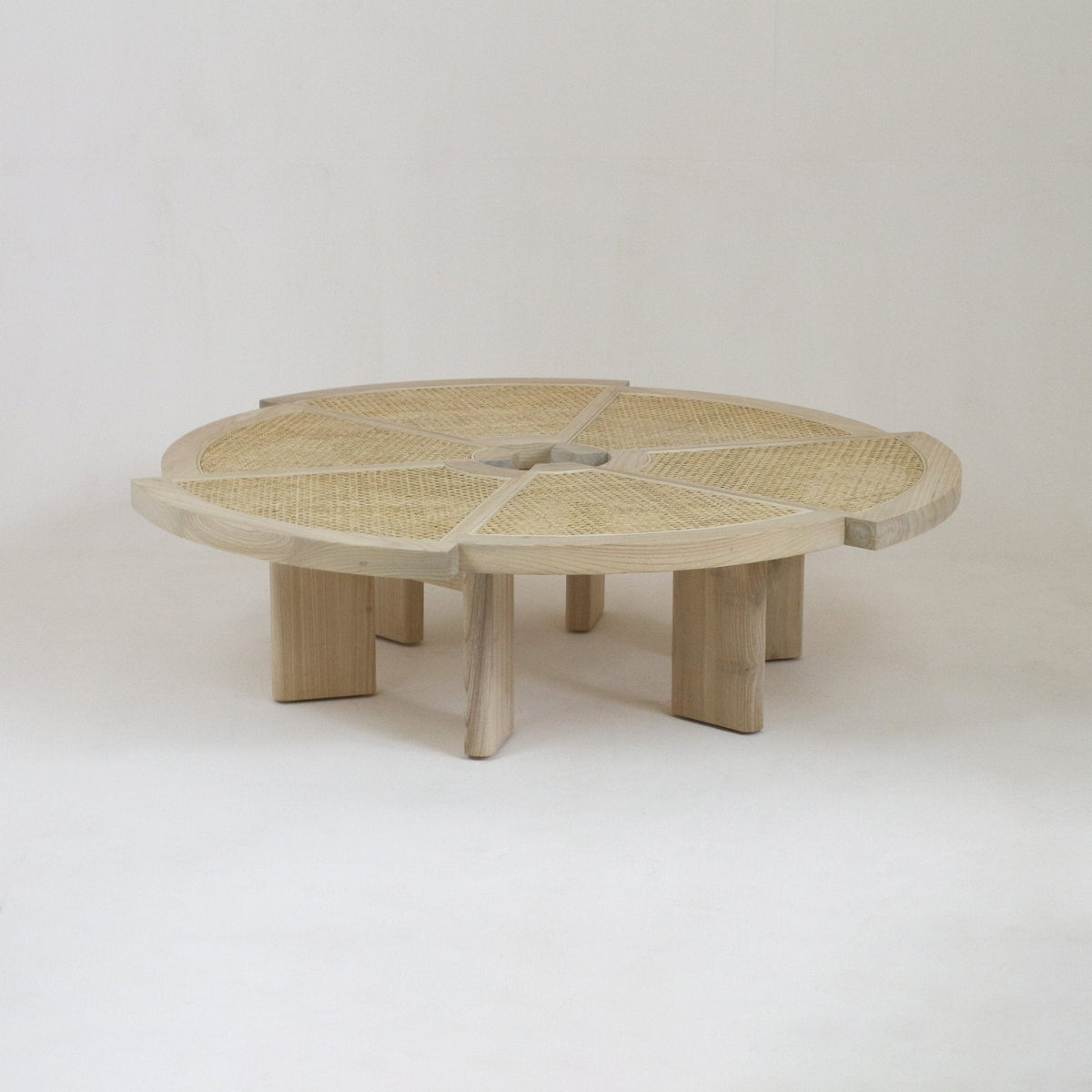 Low round coffee table in natural oak with woven rattan (cane) inset top divided into six pie-shaped panels and chunky sculpted legs, photographed on a neutral studio background.