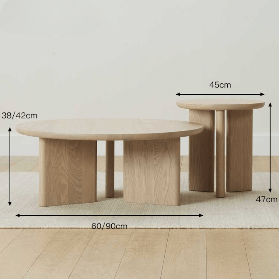 Pair of modern solid oak round coffee tables in natural oak finish with thick slab-style legs and rounded edges, shown on a light rug against a neutral wall with measurement arrows indicating sizes.