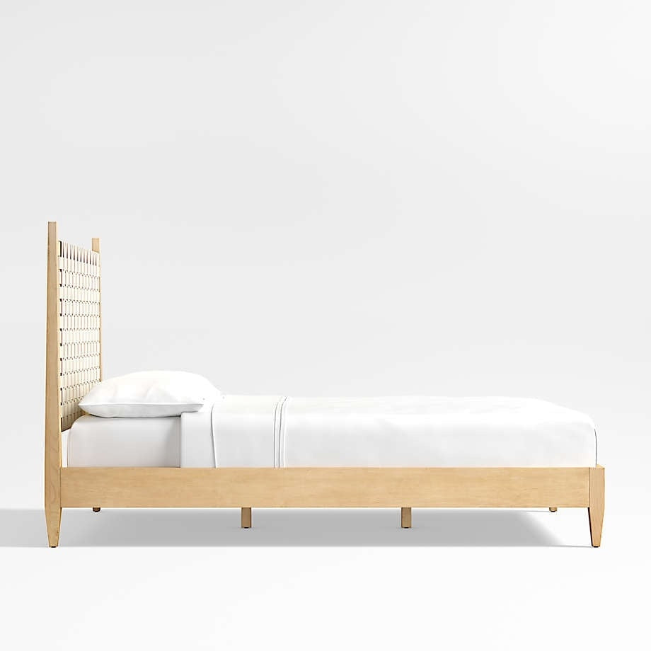 Side view of a blonde teak platform bed with a tall woven rattan headboard and low solid-wood frame, styled with crisp white sheets and a pillow in a minimalist mid-century modern design.