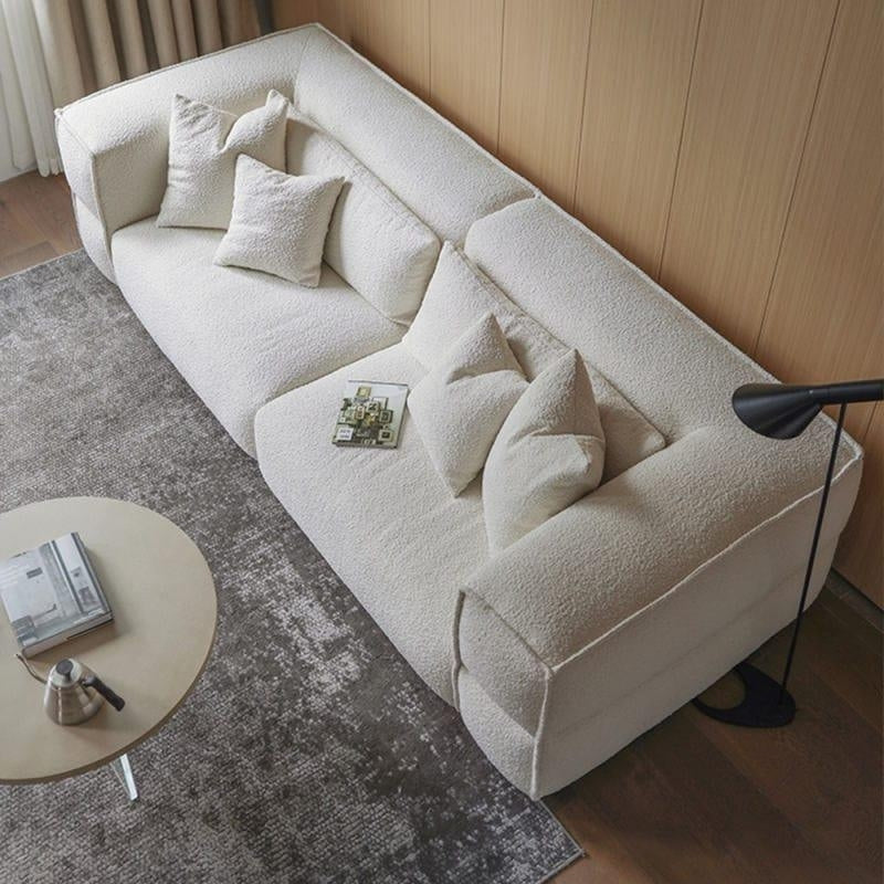 Top-down view of a modern white boucle sectional sofa with a low profile and squared arms, styled with several matching pillows, placed on a gray textured rug beside a round coffee table and a black floor lamp against a wood-paneled wall.