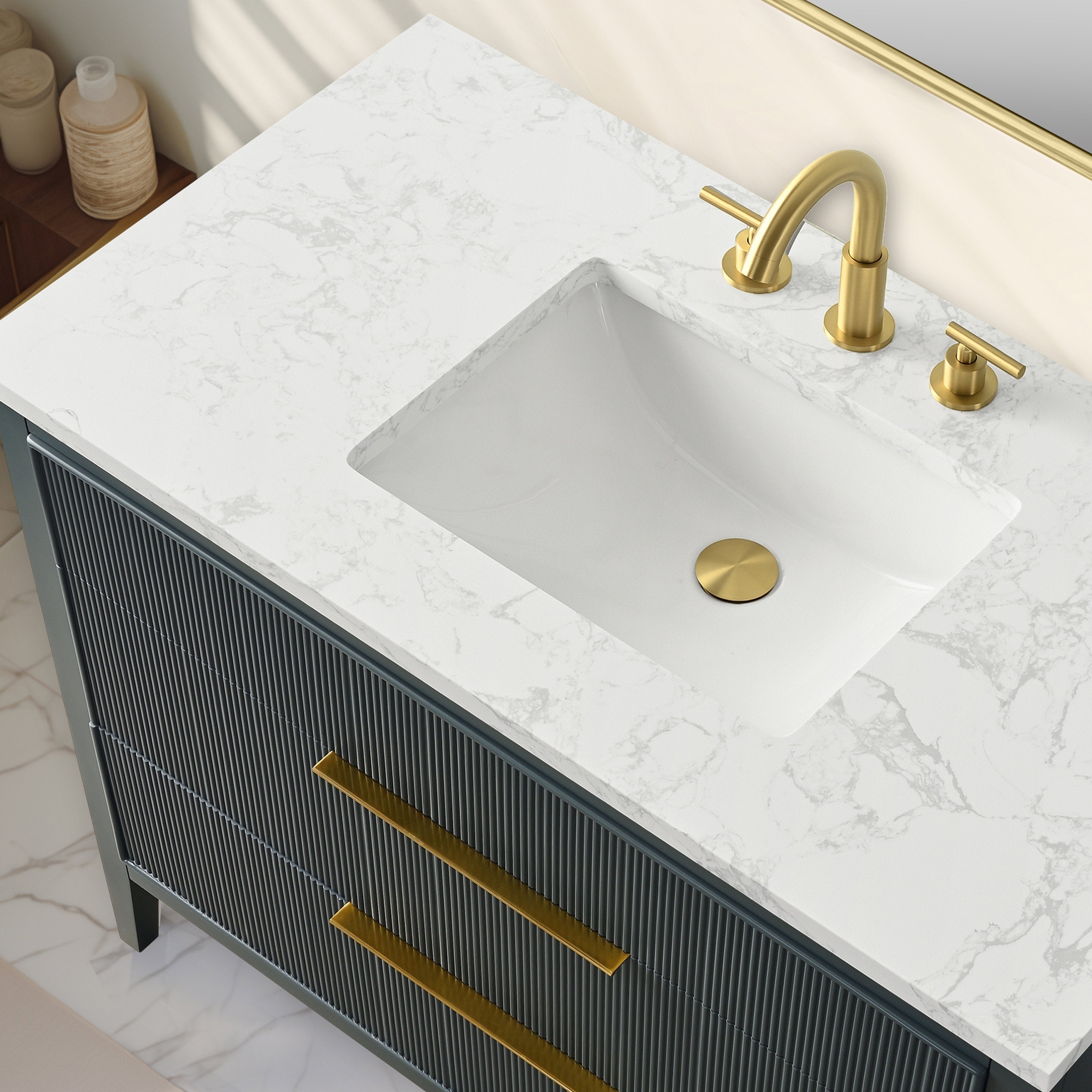Top-down view of a modern solid wood bathroom vanity with a white stone countertop with subtle gray veining, rectangular undermount ceramic sink, brushed gold faucet and drain, and long gold handles on fluted dark-wood drawers.