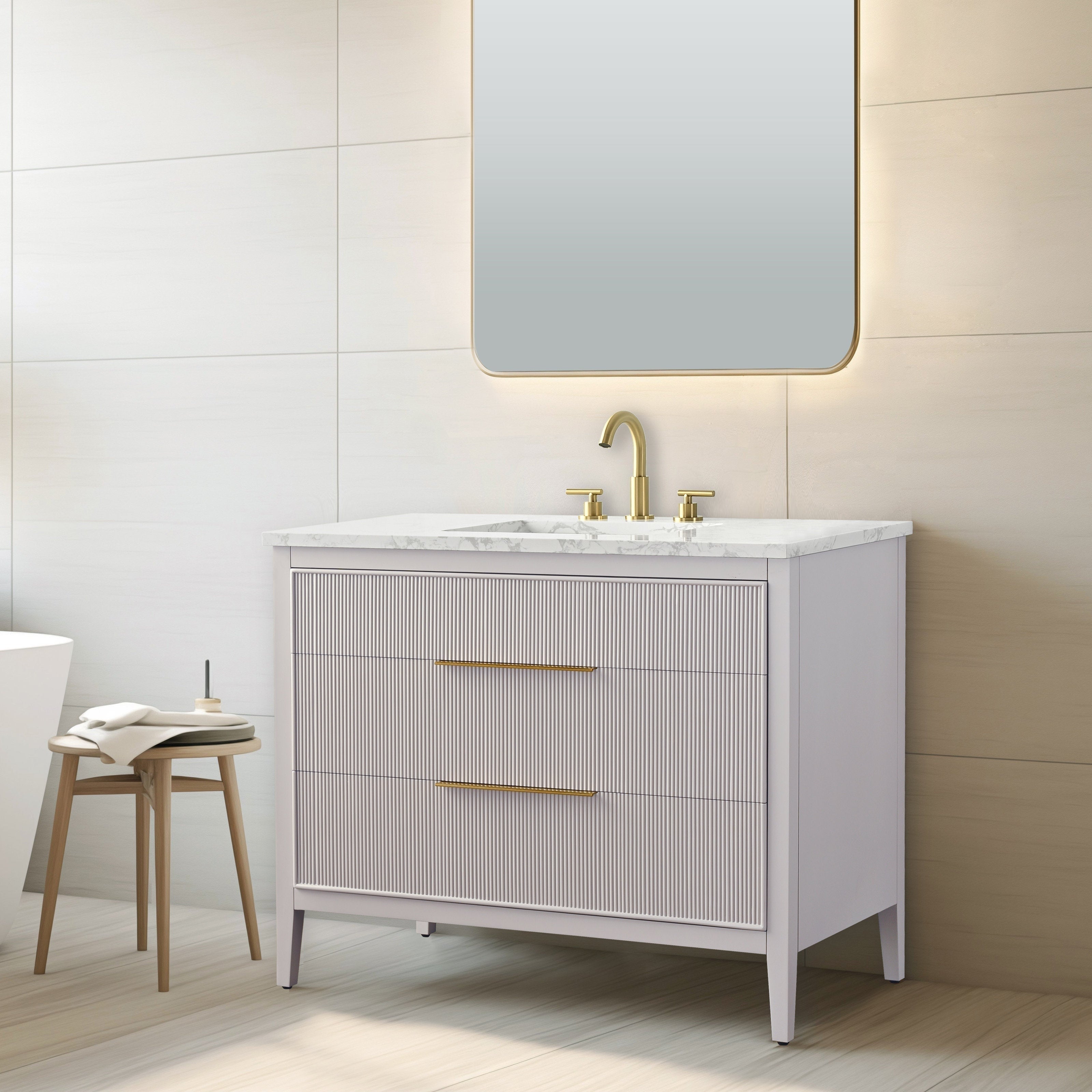 Modern freestanding single-sink bathroom vanity in stone-colored solid wood with fluted drawer fronts, white stone countertop and ceramic sink, brass faucet and long brass handles, shown under a backlit mirror with a wooden stool.