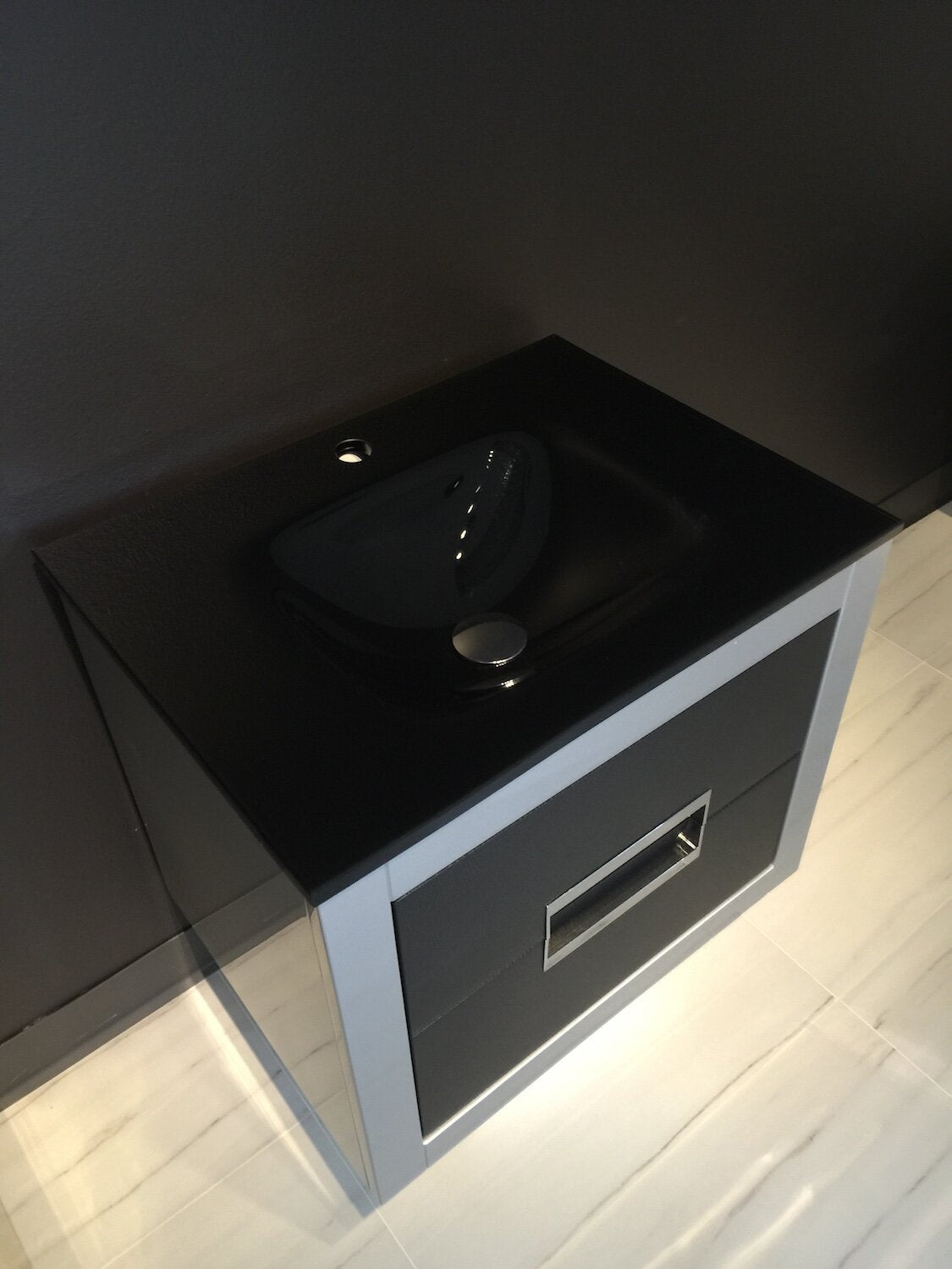 Freestanding modern single-sink bathroom vanity with a black stone top and integrated basin, solid wood frame, single gray drawer with chrome rectangular pull, photographed against a dark wall on light tile flooring.