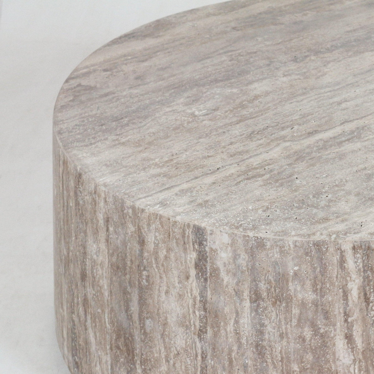 Close-up of a round natural beige travertine coffee table showing the textured stone top and vertically striated side, highlighting the material's veining and matte finish in a modern contemporary style.