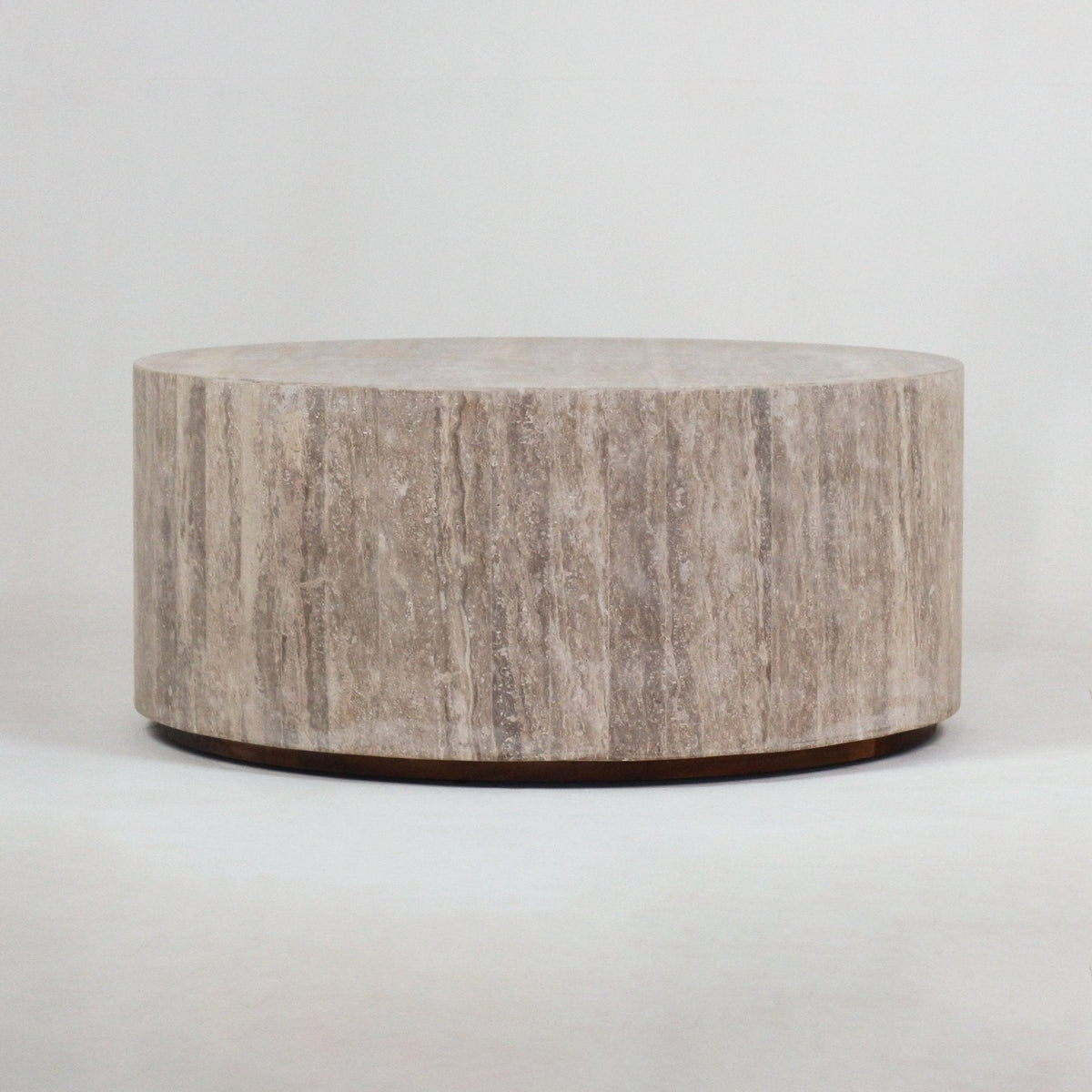 Low-profile round travertine coffee table with cylindrical drum shape, natural beige stone veining and a slim dark recessed base on a neutral studio background.