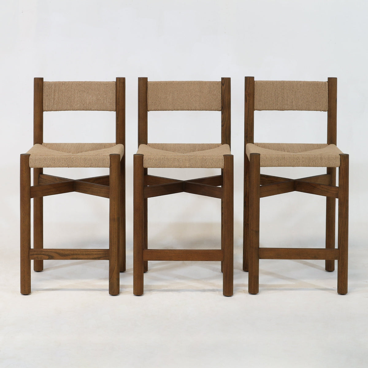 Three walnut-finished solid wood counter-height stools with natural papercord-woven seats and backs, square legs and X-shaped stretchers in a modern coastal style, photographed against a plain white background.