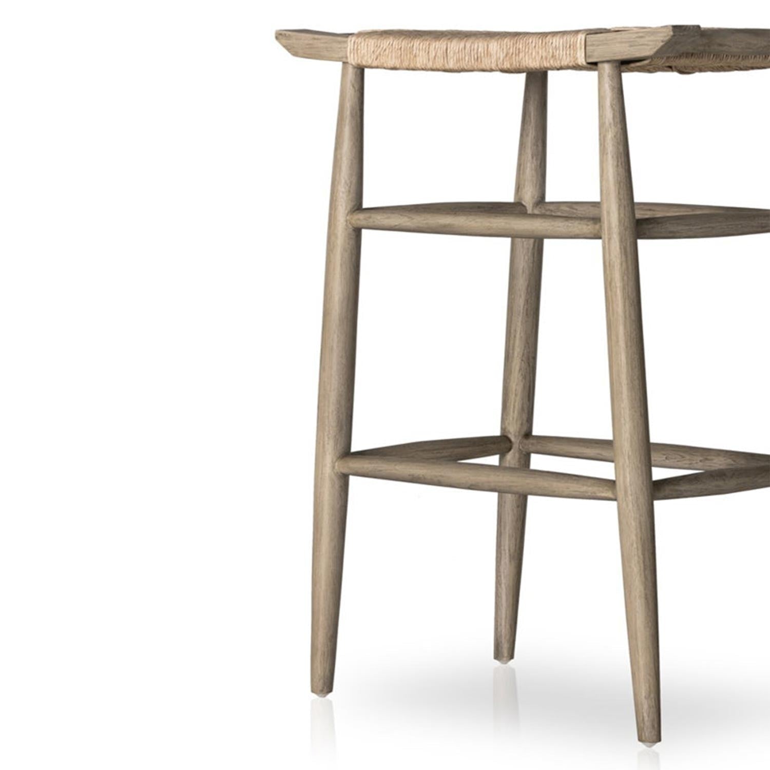 Close-up of the lower half of a natural teak backless outdoor bar stool showing tapered legs, stacked square footrest rails and woven seat edge in a coastal Scandinavian style.