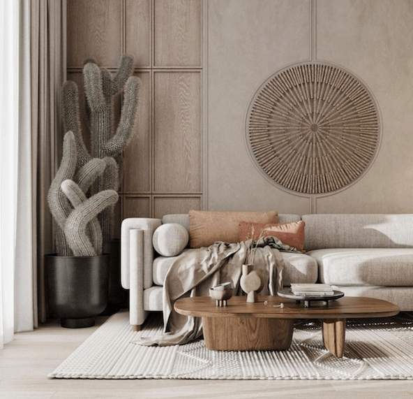 Low natural solid wood oval coffee table on a textured cream rug, styled with ceramic vases and books in front of a light beige modern sofa with rust and tan pillows, draped throw, tall potted cactus and round wooden wall art.