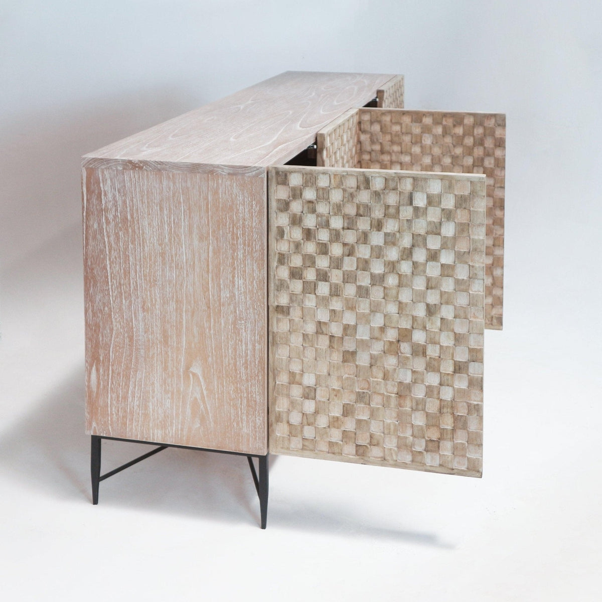 Whitewashed teak sideboard with two partially open checkerboard-carved doors, natural light wood finish and slender black metal mid-century tapered legs shown on a plain white studio background.