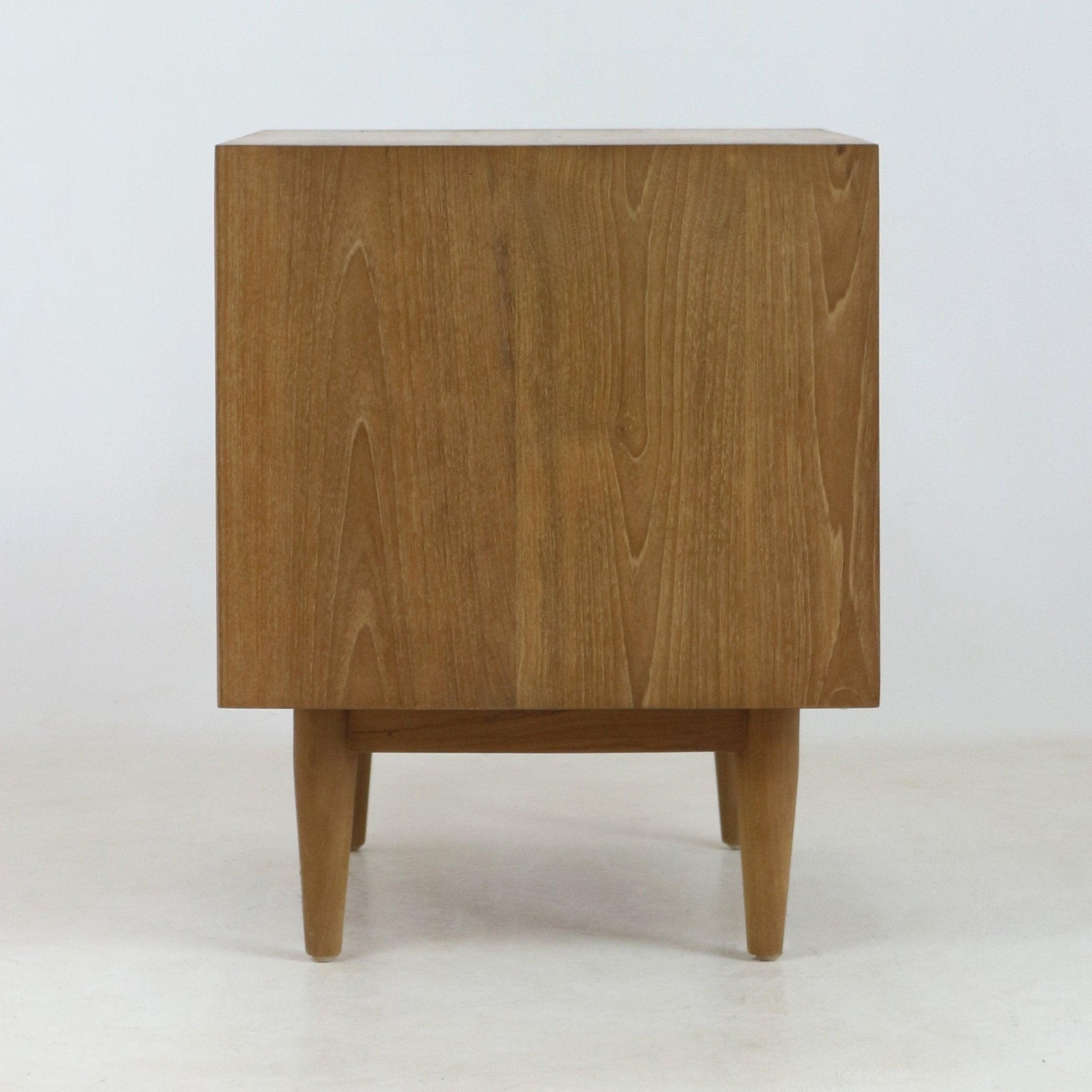 Side profile of a solid wood nightstand in natural teak finish, showing clean Mid-Century Modern lines, pronounced wood grain and tapered legs on a white studio background.