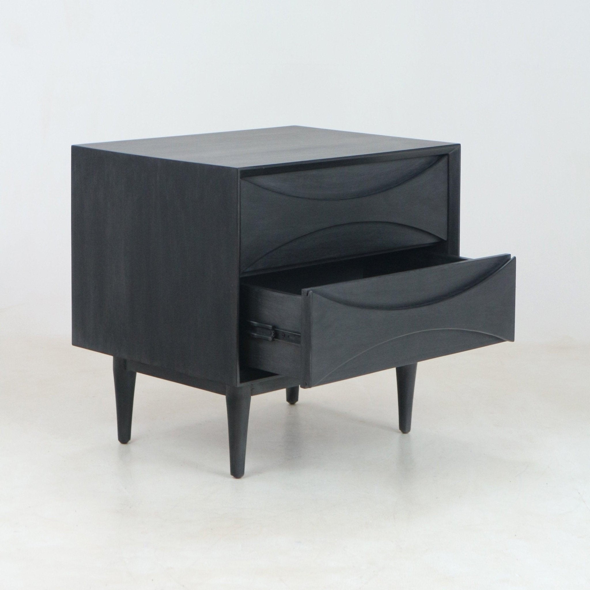 Smoked black solid wood two-drawer nightstand with sculpted curved drawer fronts, one drawer open and tapered mid-century modern legs set on a light studio floor.