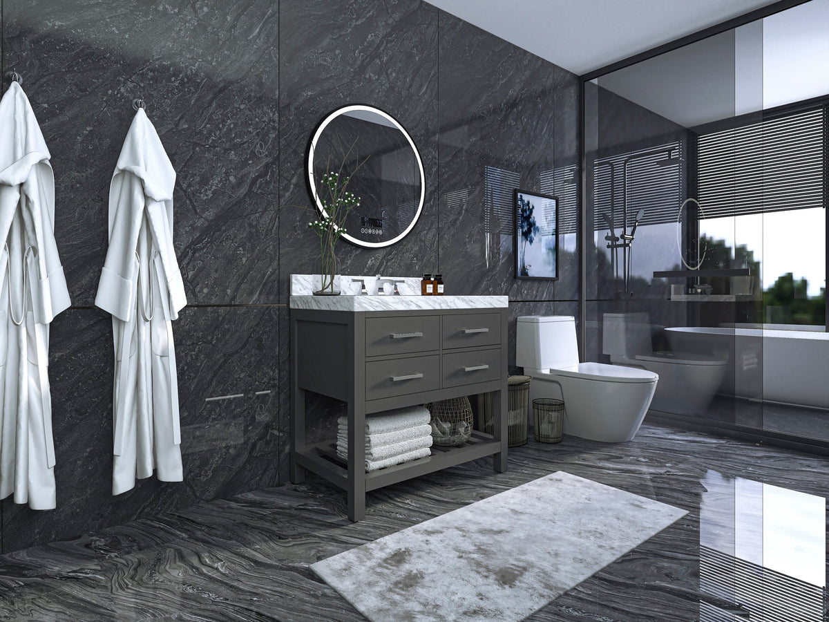 Modern solid wood gray bathroom vanity with white stone marble top, round mirror, chrome faucet and open lower shelf with folded towels, set against dark stone walls and a glass shower enclosure.