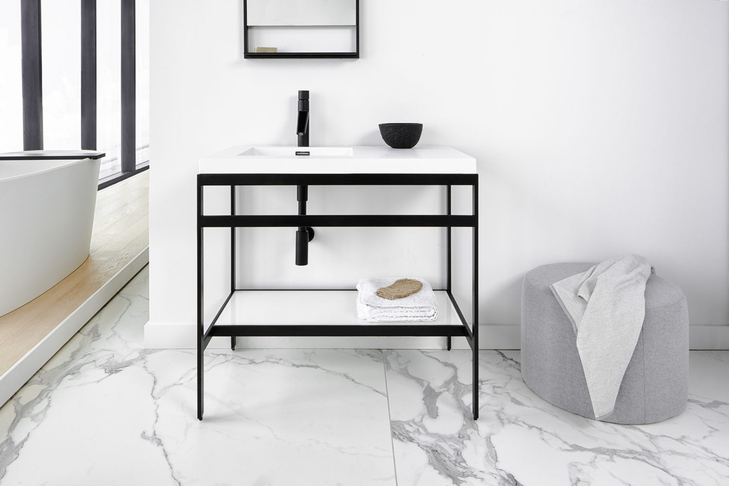 Modern freestanding bathroom vanity with a white stone top and integrated ceramic sink, supported by a slim black metal frame with open shelf holding folded towels, styled in a minimalist marble-floored bathroom.