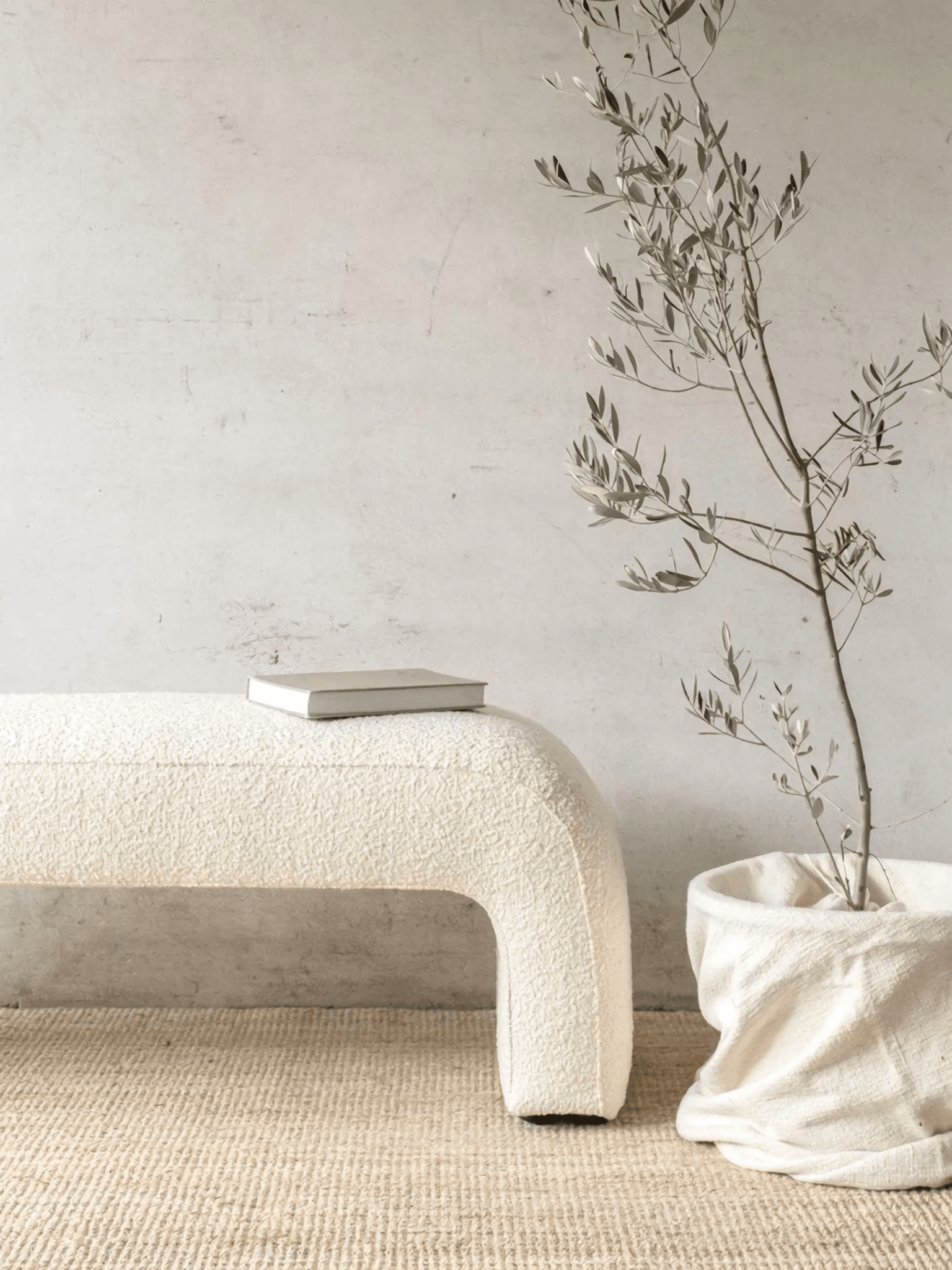 Cream boucle upholstered arched bench on a woven jute rug, styled with a closed neutral book on top and a potted branch in a soft fabric basket against a textured off-white wall.