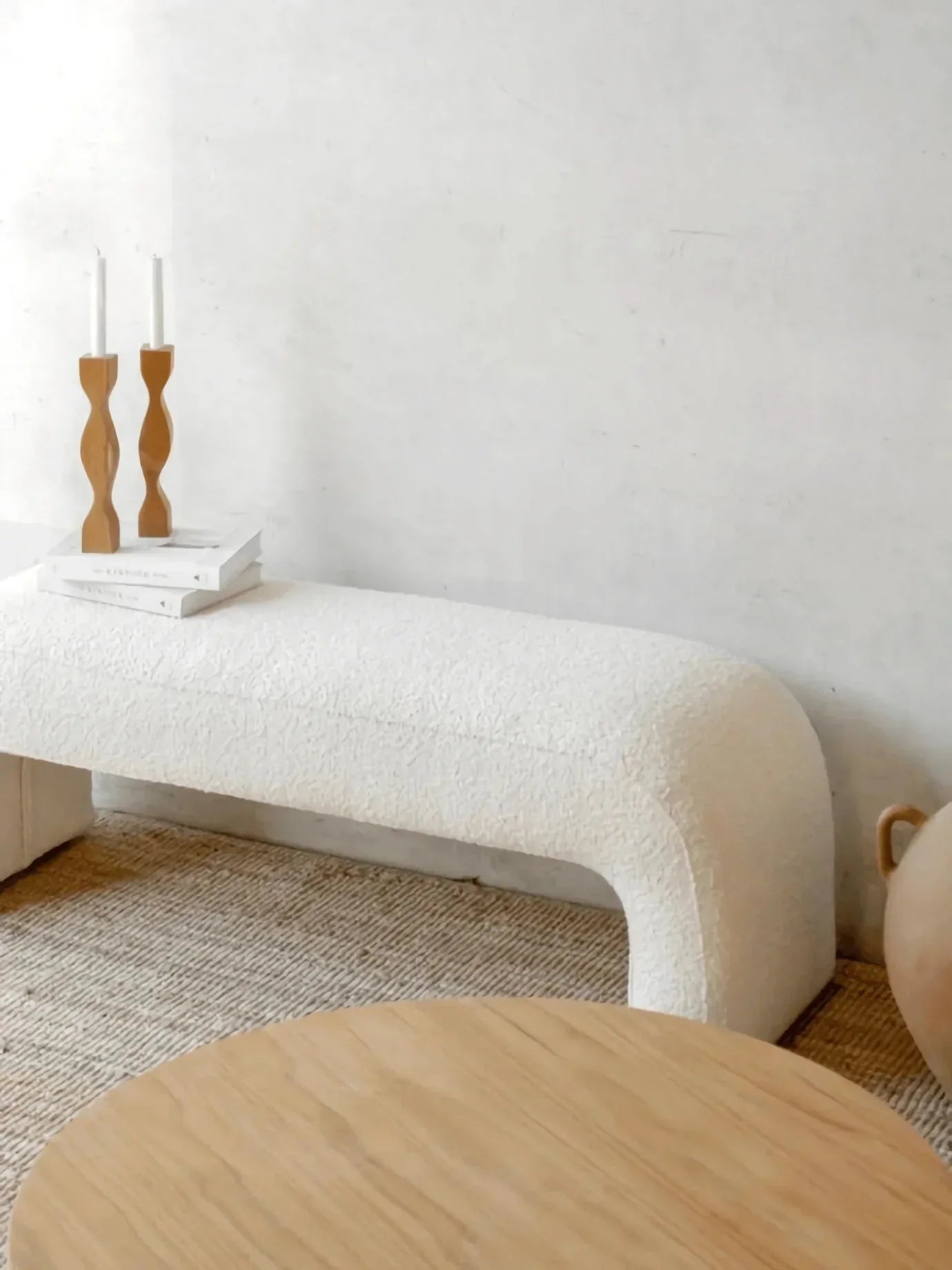 Backless 59-inch boucle upholstered bench in soft white with rounded U-shaped legs, styled on a woven jute rug with a round oak coffee table, stacked white books and carved wooden candlesticks in a coastal-contemporary setting.