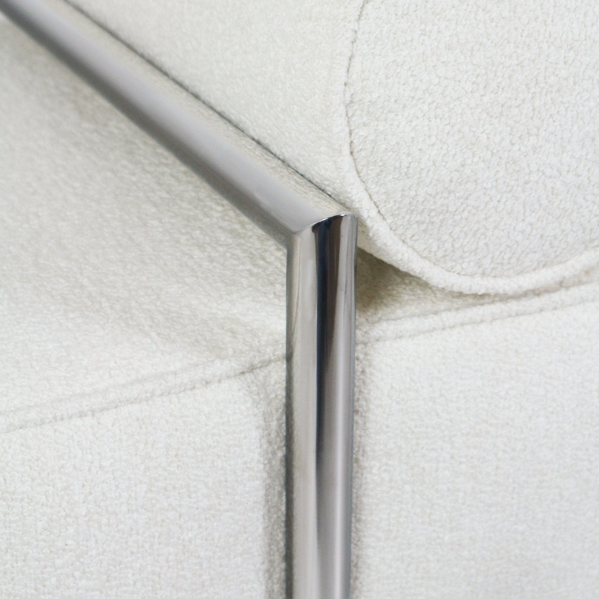 Close-up of a polished stainless-steel leg and angled rail against cream boucle upholstery with a rounded bolster on an end-of-bed bench.