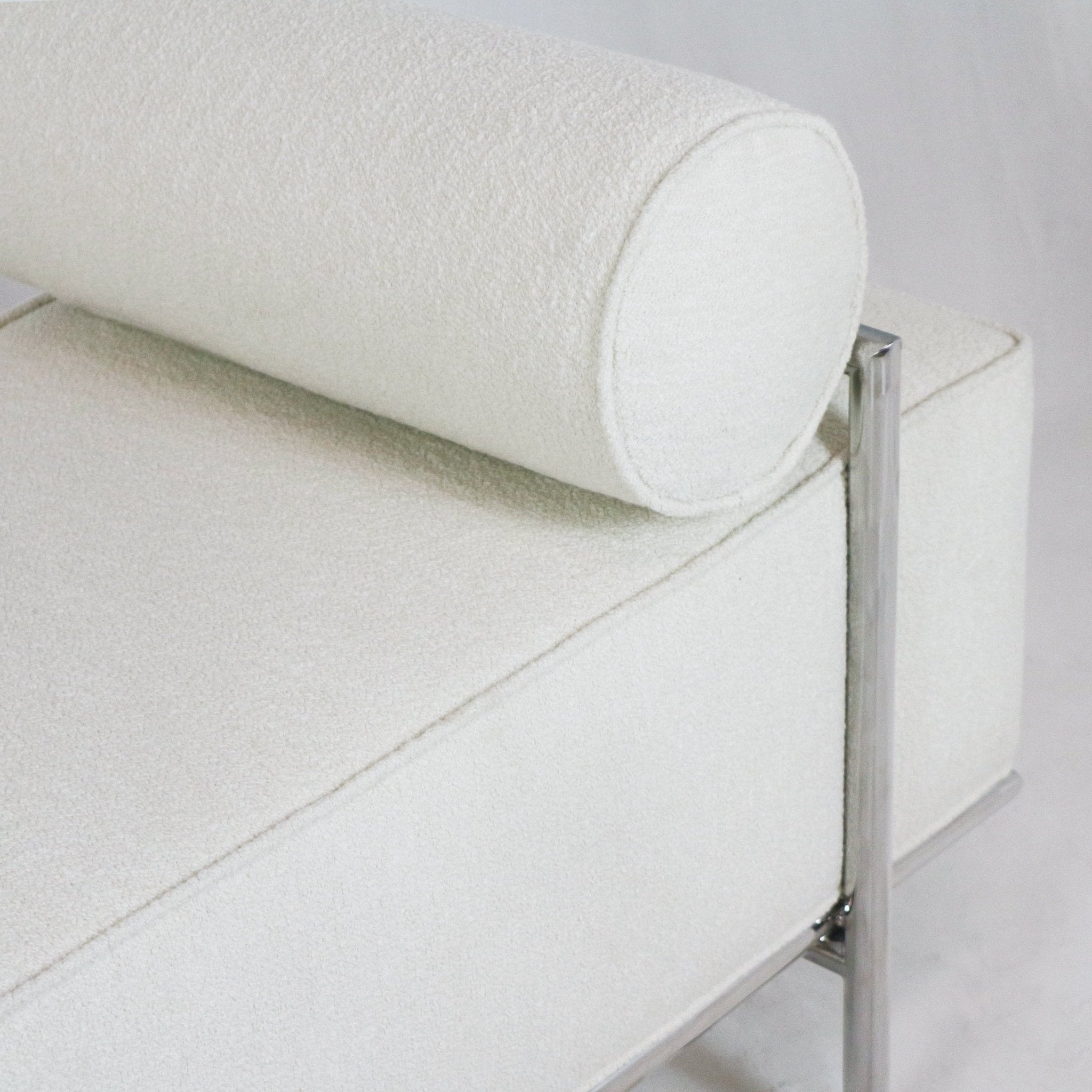 Close-up of a cream boucle-upholstered end-of-bed bench corner showing a cylindrical bolster pillow, stitched seat seam, and polished metal leg and frame.
