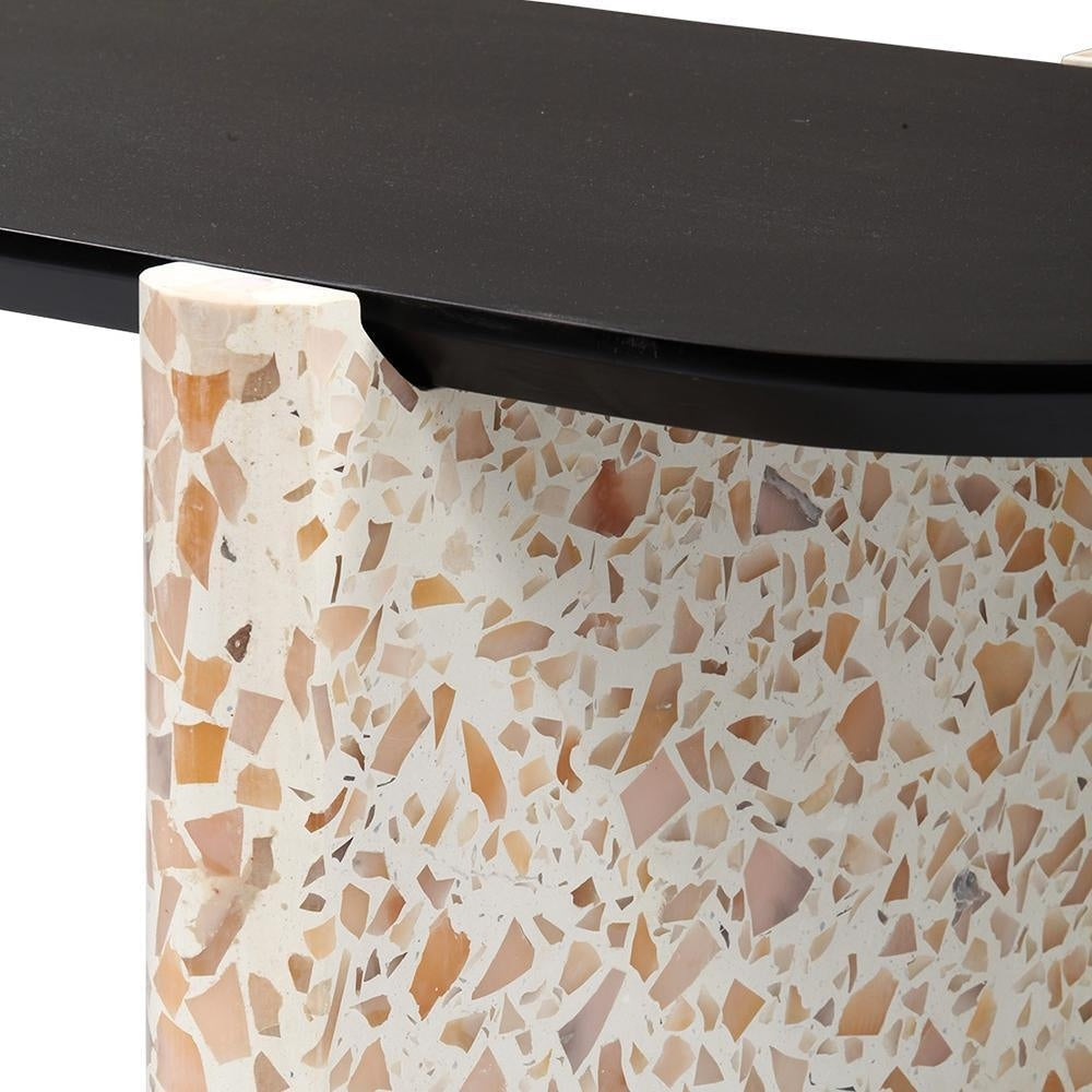 Close-up of a modern coffee table showing a black wooden tabletop edge resting on a rounded terrazzo stone base with a cream background and warm beige, pink and amber chips.