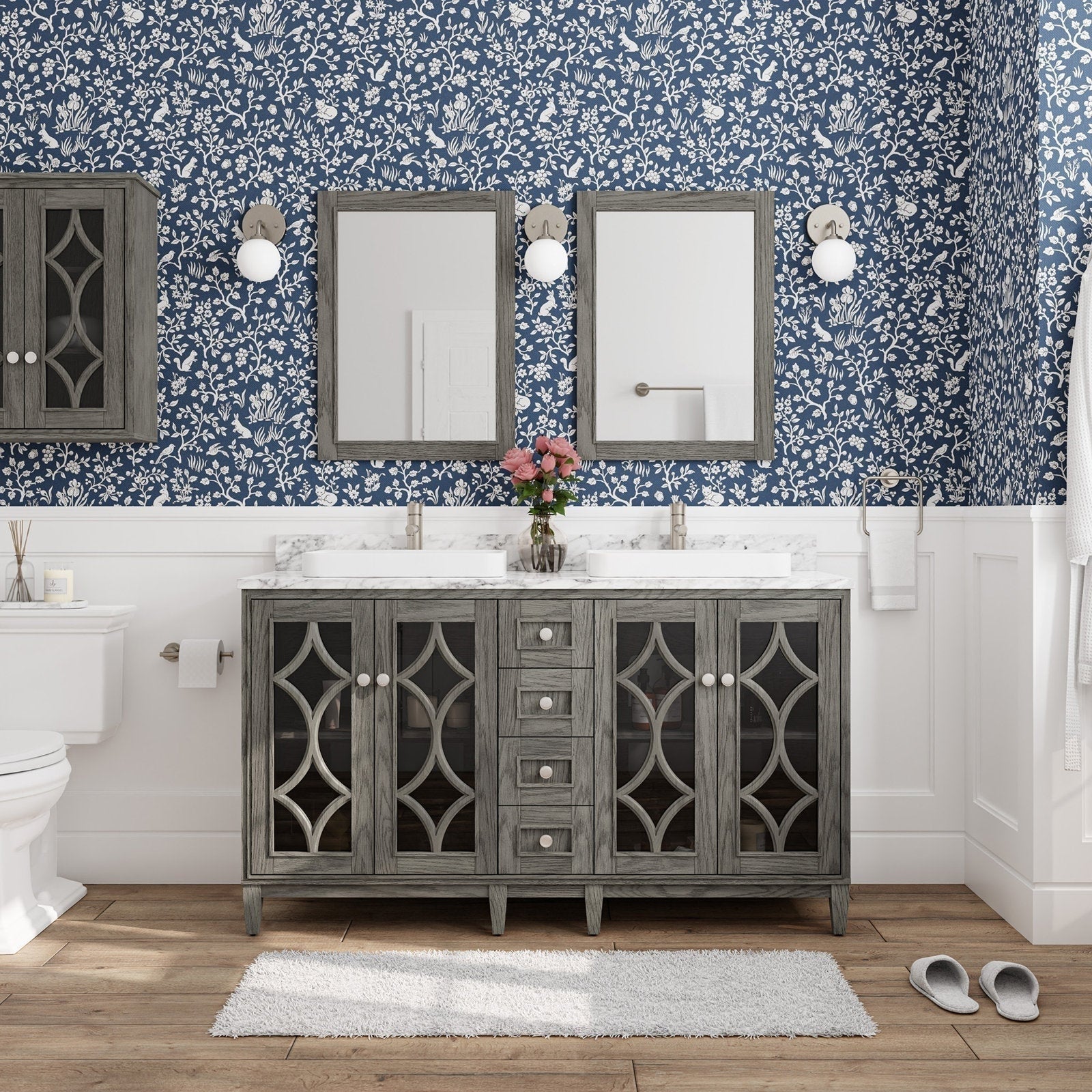 Freestanding solid-wood double bathroom vanity in weathered gray with a stone top, two rectangular above-counter sinks and brushed nickel faucets, glass-front lattice cabinet doors, center drawers, and matching mirrors against blue floral wallpaper.
