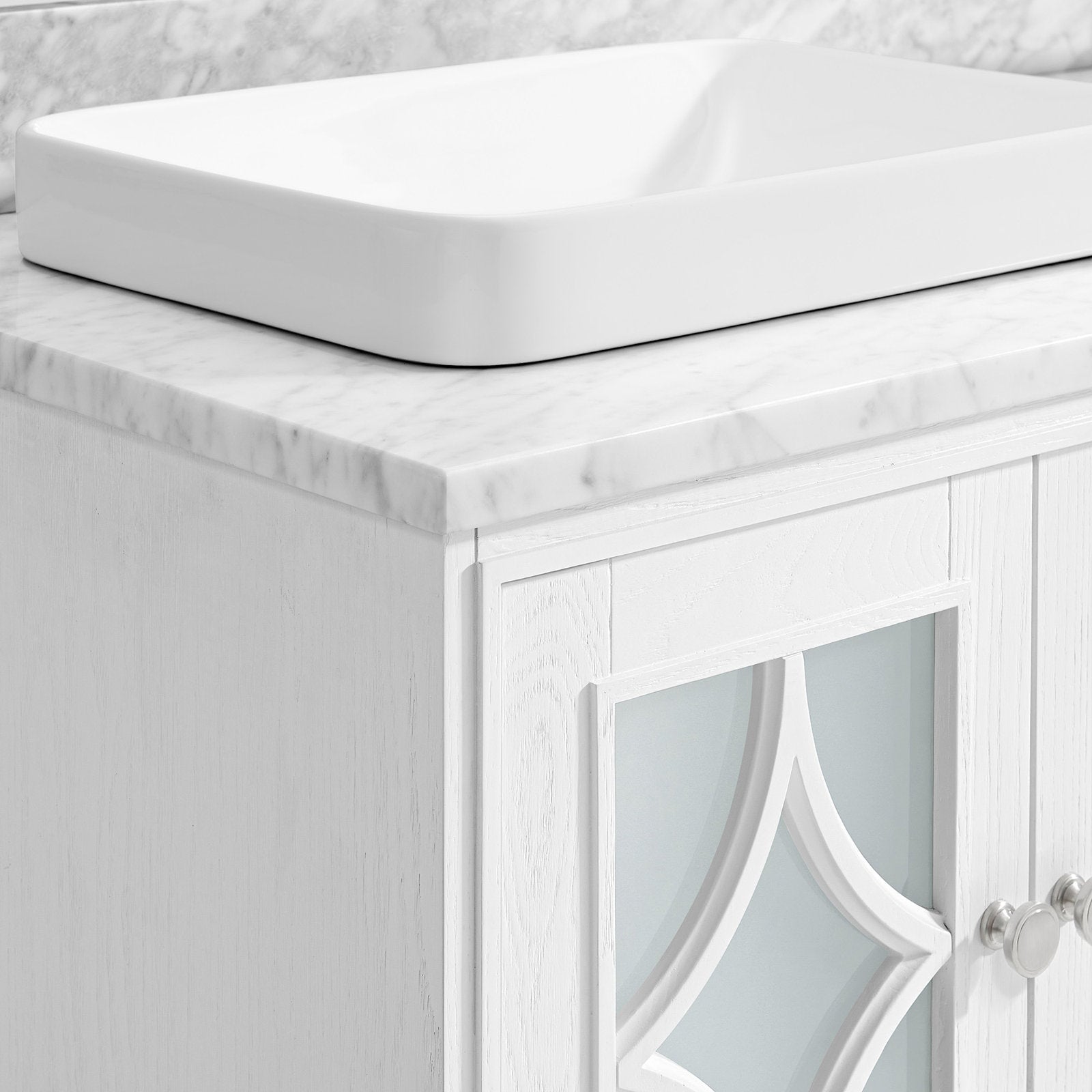 Close-up of a stone marble-look countertop with a white rectangular ceramic vessel sink and the corner of a white solid-wood vanity door featuring frosted decorative glass and round metal knobs.