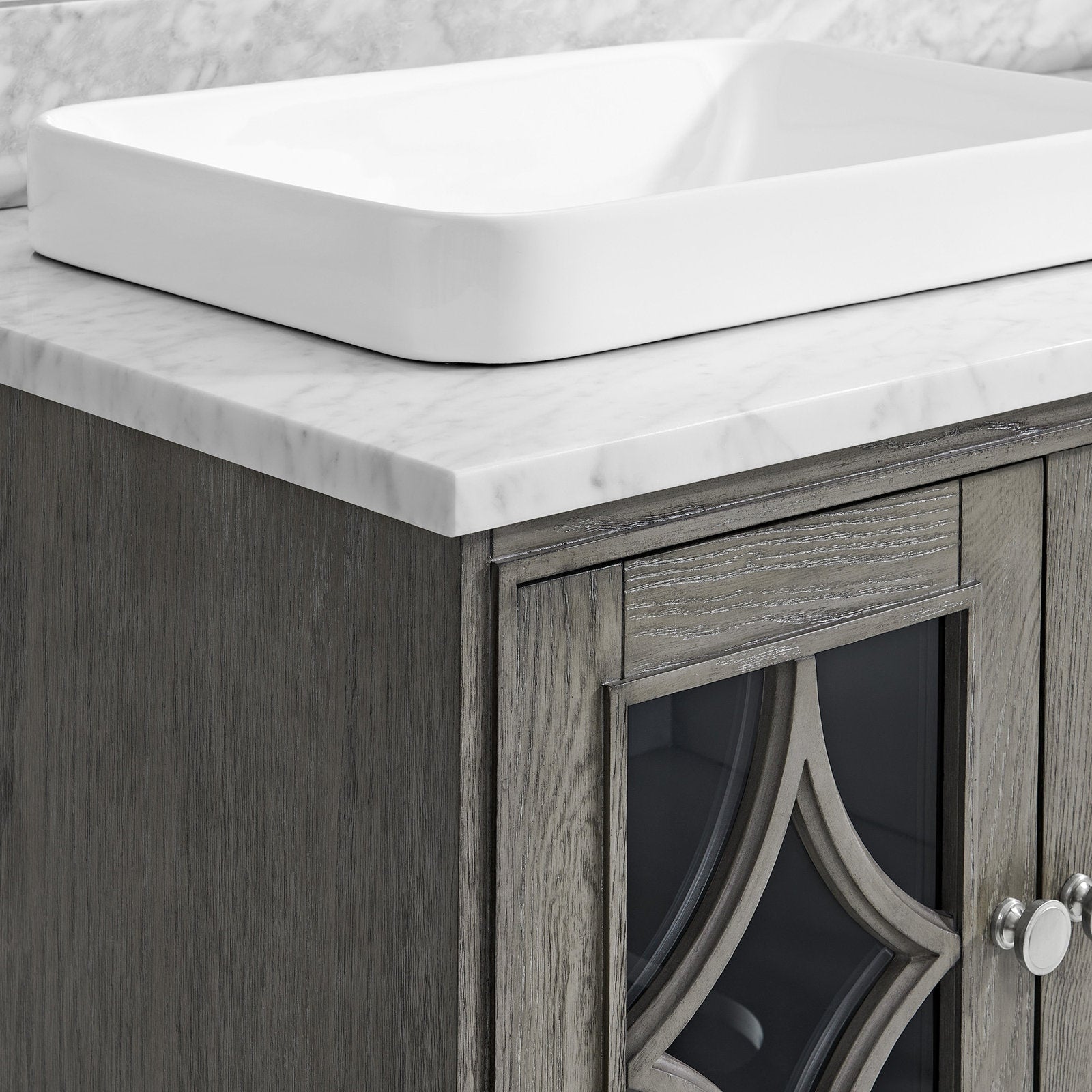 Close-up of a white rectangular ceramic vessel sink on a white stone countertop with subtle gray veining, mounted atop a gray-stained solid wood vanity with a decorative glass-front door and round metal knob.