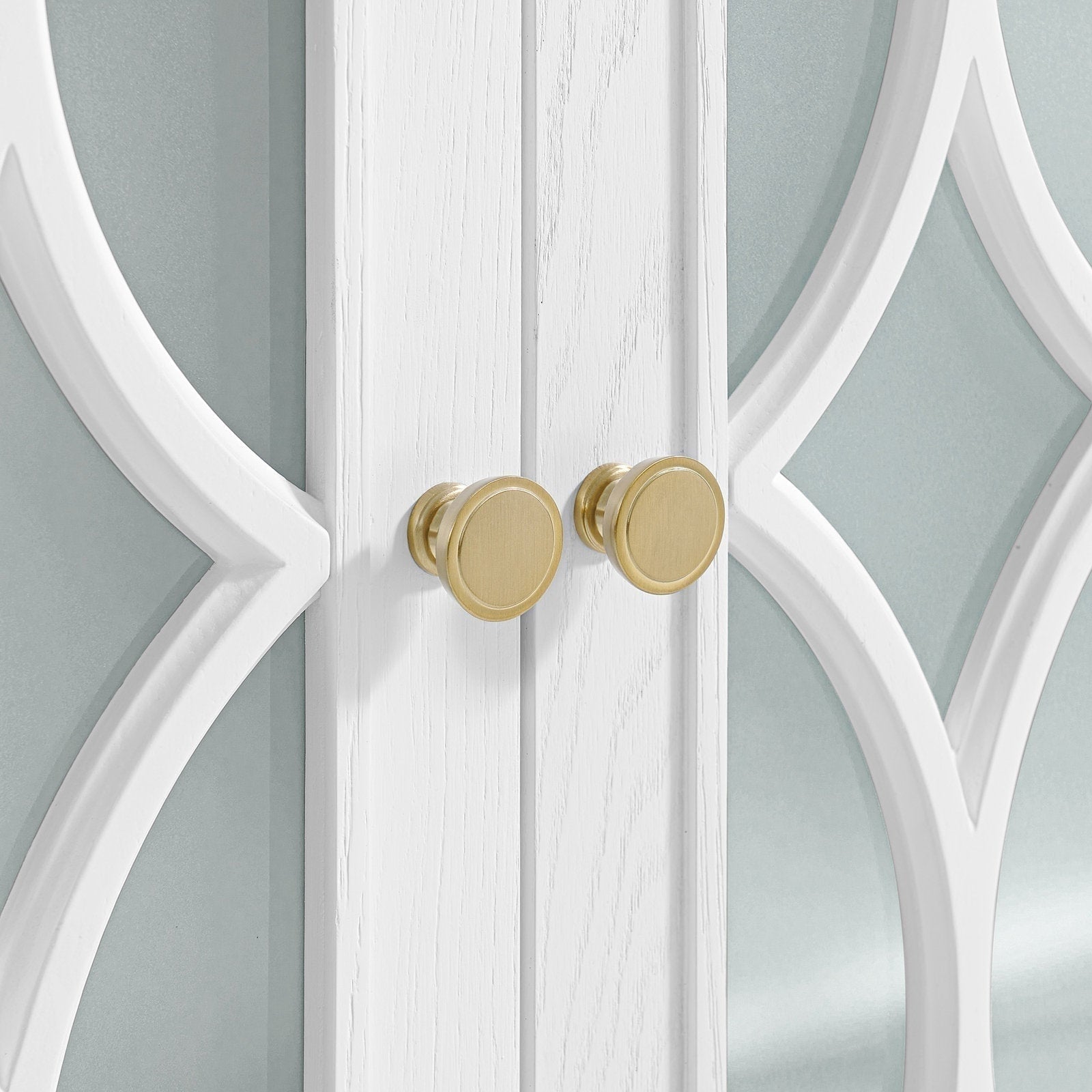 Close-up of white solid-wood cabinet doors with decorative white fretwork over frosted stone-colored glass, showing two round brushed brass knobs centered on the doors' meeting stile.