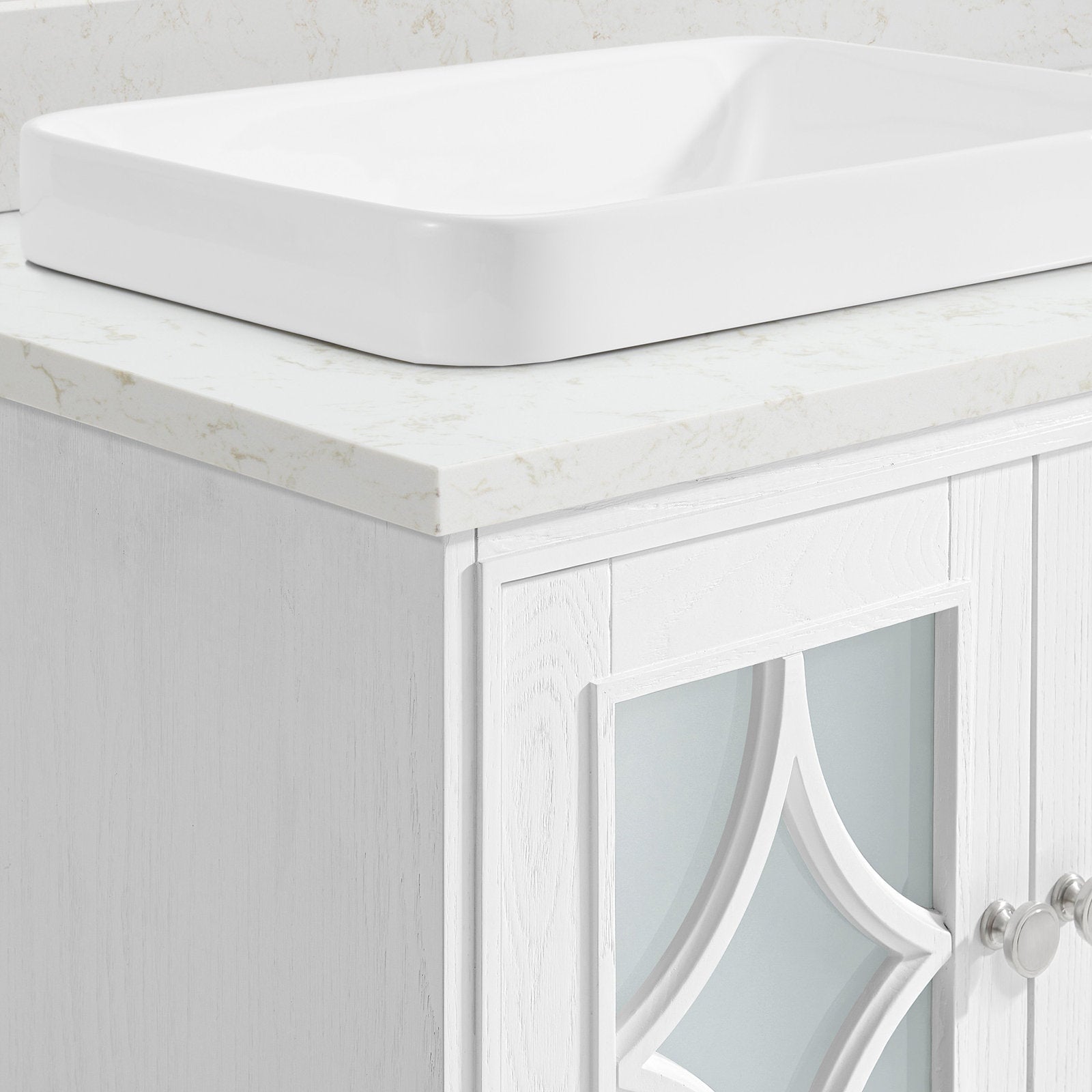 Close-up of a stone-look countertop with a white ceramic vessel sink resting on a white painted solid-wood vanity featuring a frosted decorative glass panel and silver round knobs.