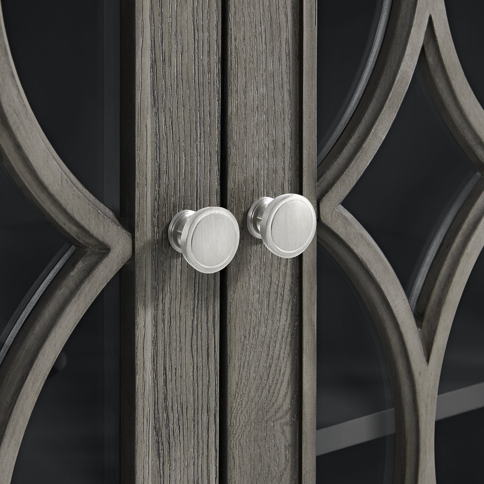 Close-up of stone-colored solid wood vanity doors with decorative curved mullions and two round brushed-metal knobs over dark glass panels, showing textured wood grain and modern hardware.