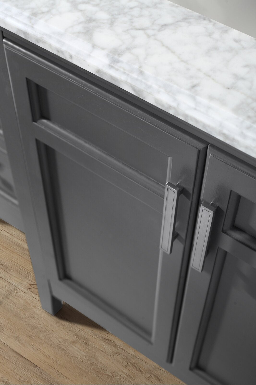 Close-up of a modern bathroom vanity showing a white stone marble countertop and gray solid wood cabinet with recessed shaker-style panels and rectangular metal handles on a light wood floor.