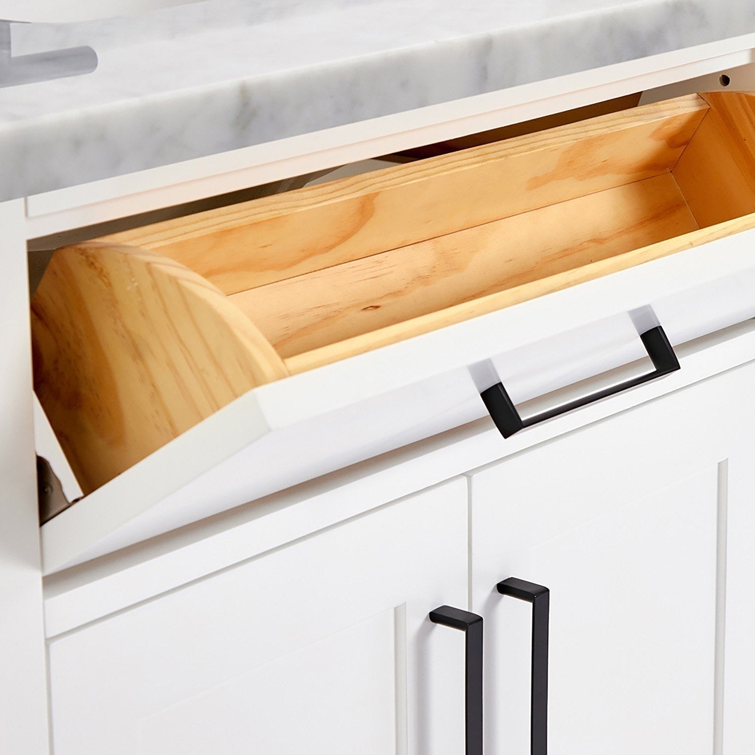 Close-up of a white bathroom vanity tilt-out compartment with a natural wood interior, white quartz countertop edge, and matte-black rectangular drawer and door handles.