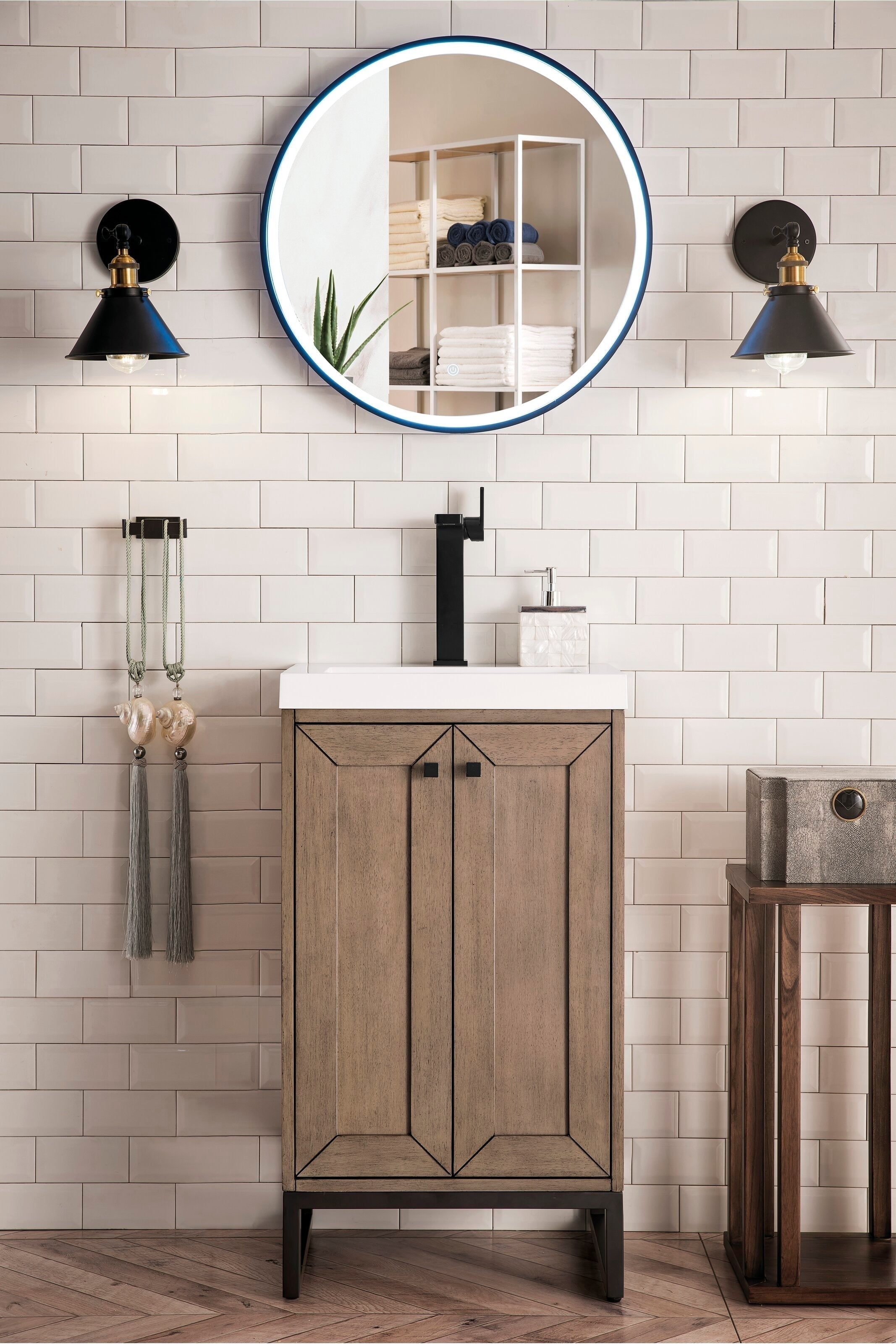 Freestanding single-sink bathroom vanity in stone-finish solid wood with a white ceramic top and matte black tall faucet, shown beneath a round LED mirror and flanked by black wall sconces on subway tile.