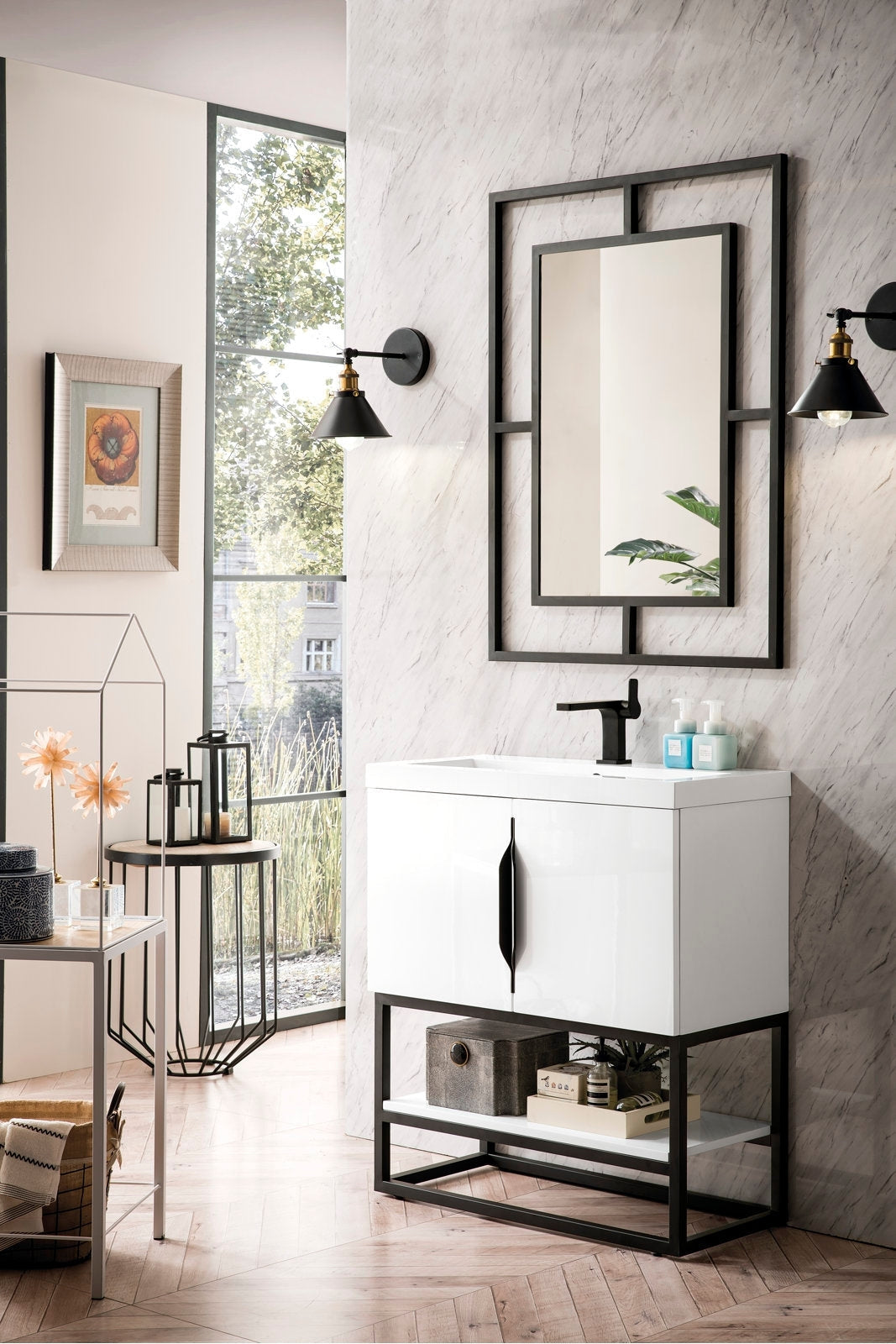 Modern white single-sink bathroom vanity with a stone top, black metal base and vertical black pull, matte black faucet, geometric black-framed mirror and wall sconces over a marble-effect wall; open lower shelf holds a storage box and tray.