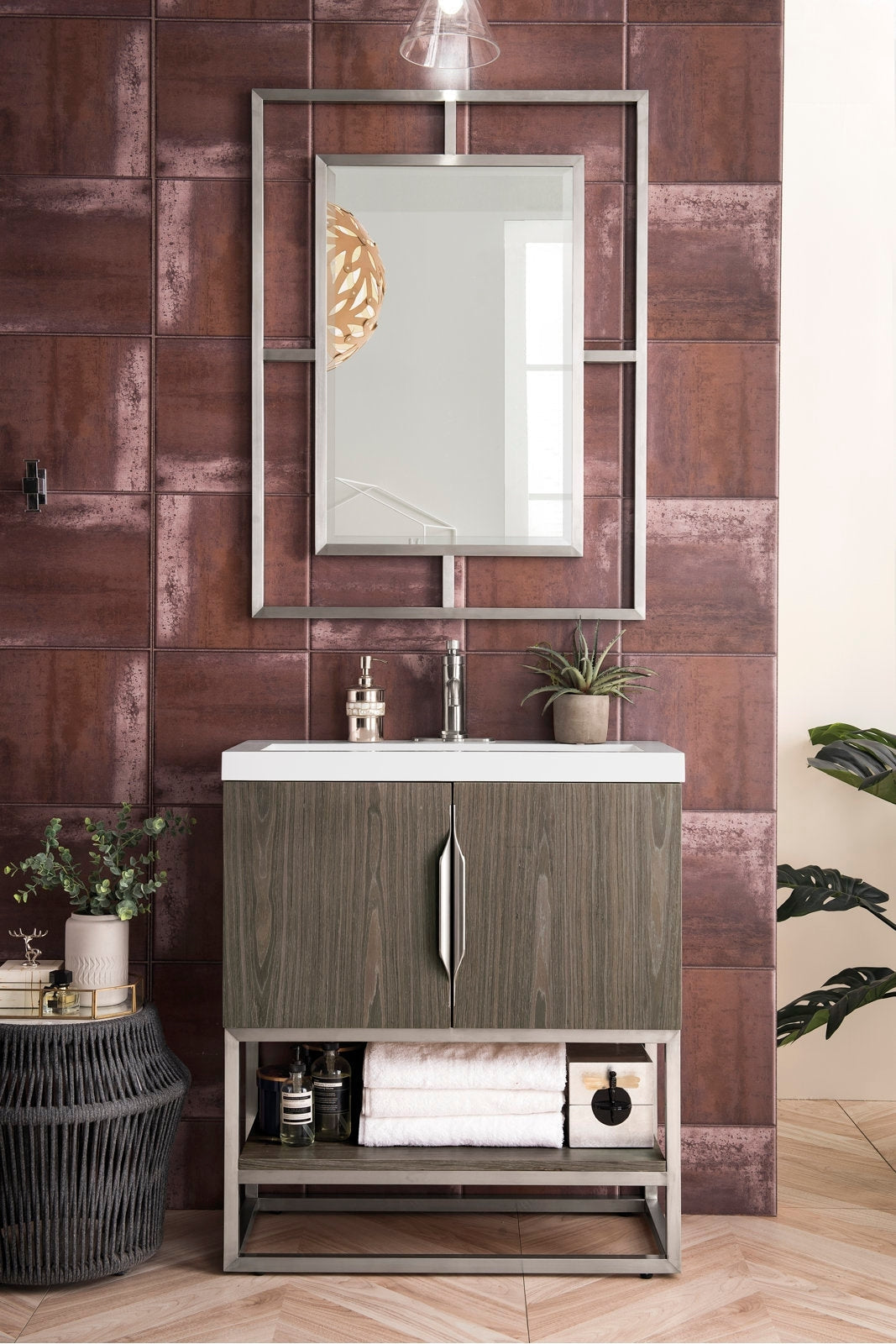 Freestanding modern single-sink bathroom vanity with a white stone top, solid wood stone-gray cabinet, metal base and chrome faucet, framed mirror above, and open lower shelf holding folded towels and bath accessories.