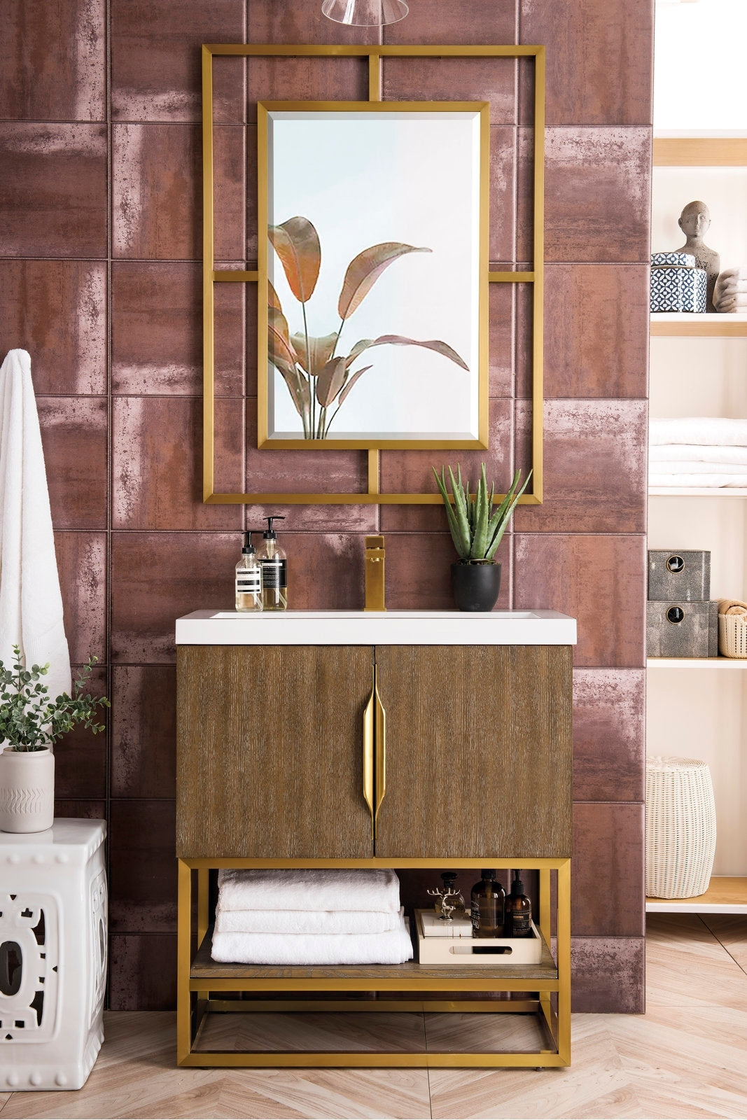 Contemporary solid wood bathroom vanity in a stone finish with a white stone top and integrated ceramic sink, gold-tone faucet and hardware, open lower shelf with folded towels, and a gold-framed mirror on textured tile.