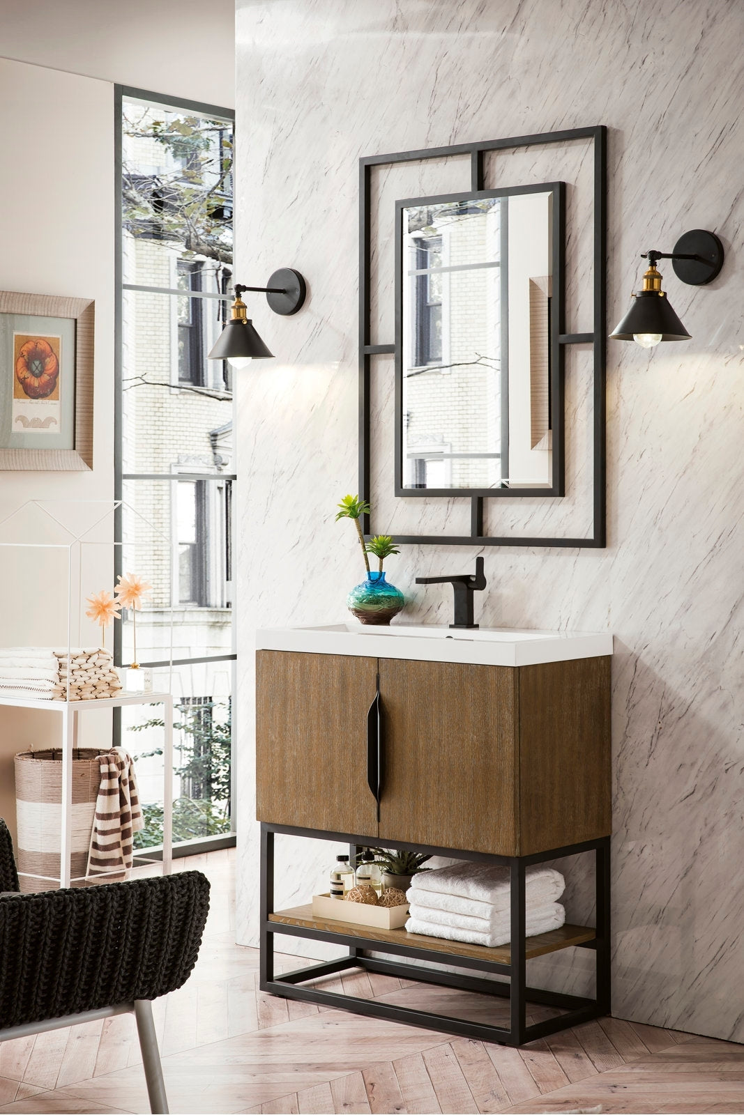Freestanding modern single-sink vanity with a stone top and solid wood stone-colored cabinet on a black metal base, black faucet and handle, shown with a black-framed mirror, two wall sconces, towels and a decorative vase.