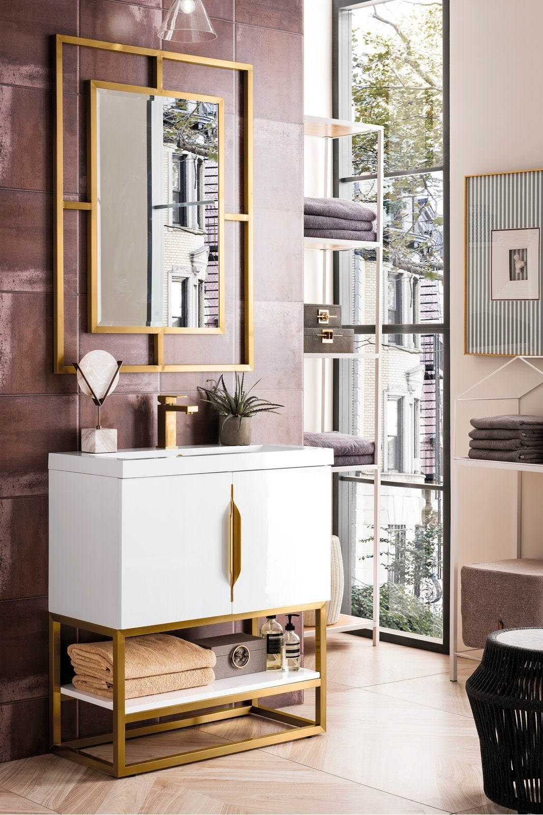 Freestanding modern white single-sink bathroom vanity with a stone top, gold metal base and matching gold handle, styled with folded towels, toiletries and a geometric gold-framed mirror by a tall window.