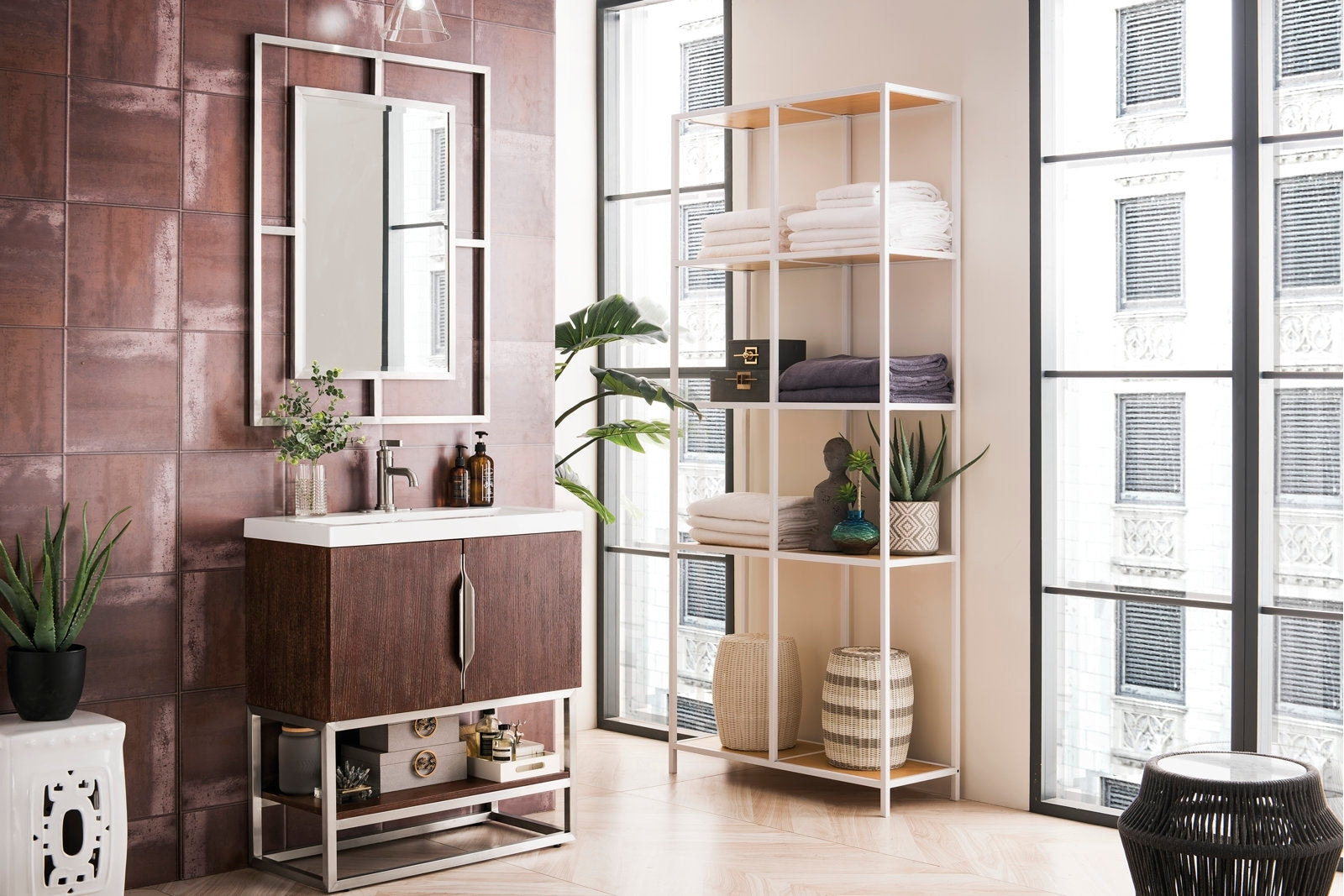Freestanding modern solid wood single-sink vanity in dark wood with a white stone top, metal base and framed mirror, styled with faucet and bottles; shown next to open shelving with towels, baskets and plants.